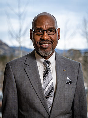 A man wearing glasses, a gray suit, a patterned tie, and a pocket square smiles at the camera with a blurred natural background.