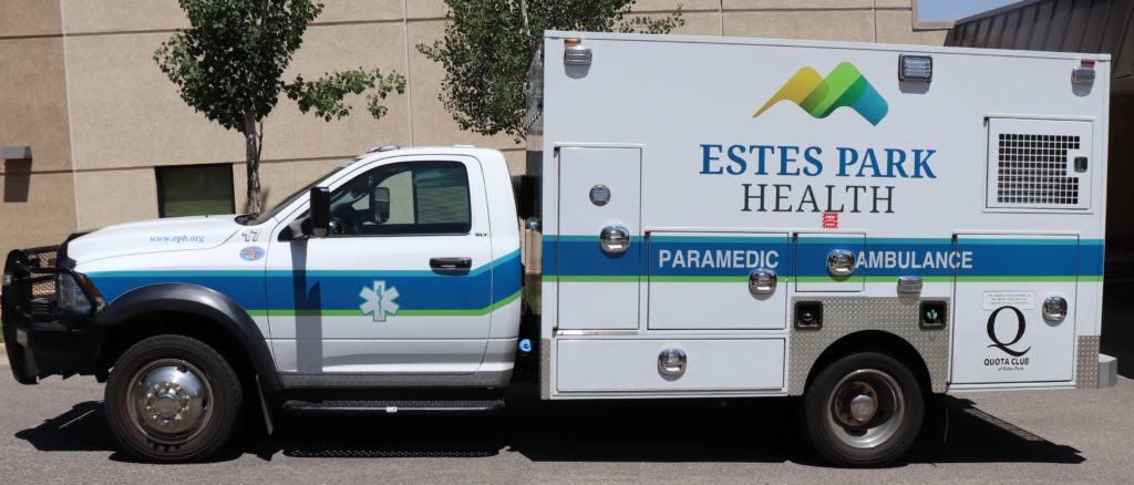 A white paramedic ambulance truck labeled "Estes Park Health" with a blue and green stripe is parked in front of a building. Trees and a portion of the building are visible in the background.