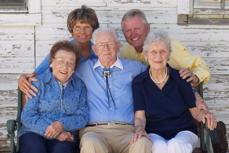 A group of six older adults poses in front of a weathered wooden building, three seated and three standing behind them, all smiling at the camera.