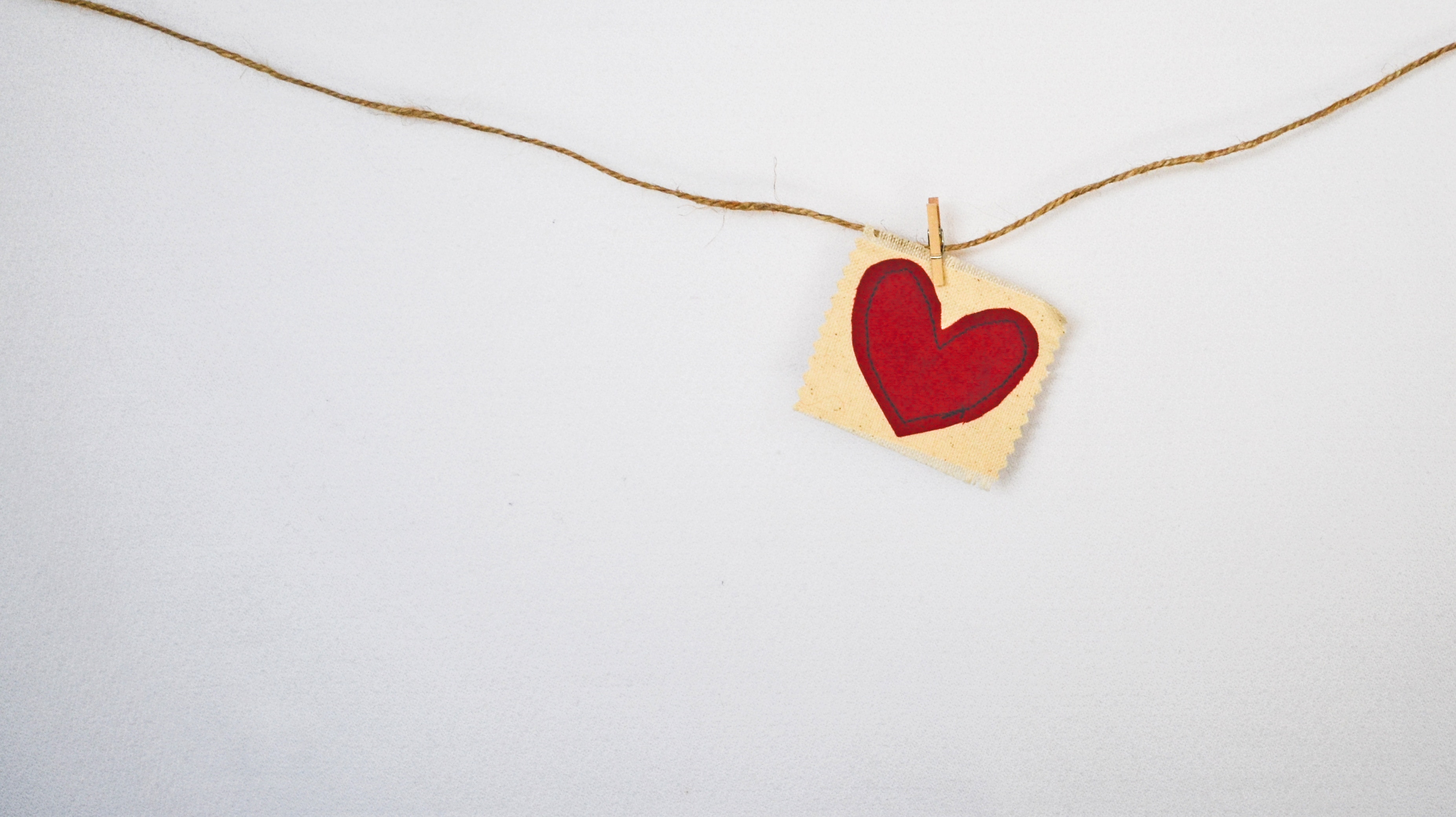 A piece of twine is stretched across a light background, holding a small square paper with a red heart on it, fastened by a mini wooden clothespin.