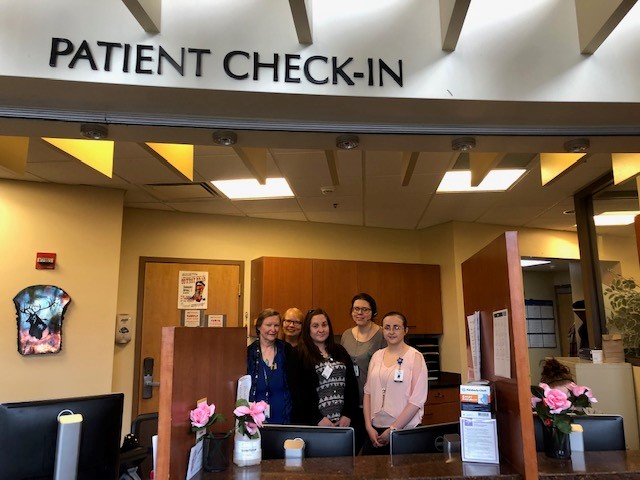 Group of five people standing behind the counter at a patient check-in area inside a clinic or hospital, with a sign above them reading 