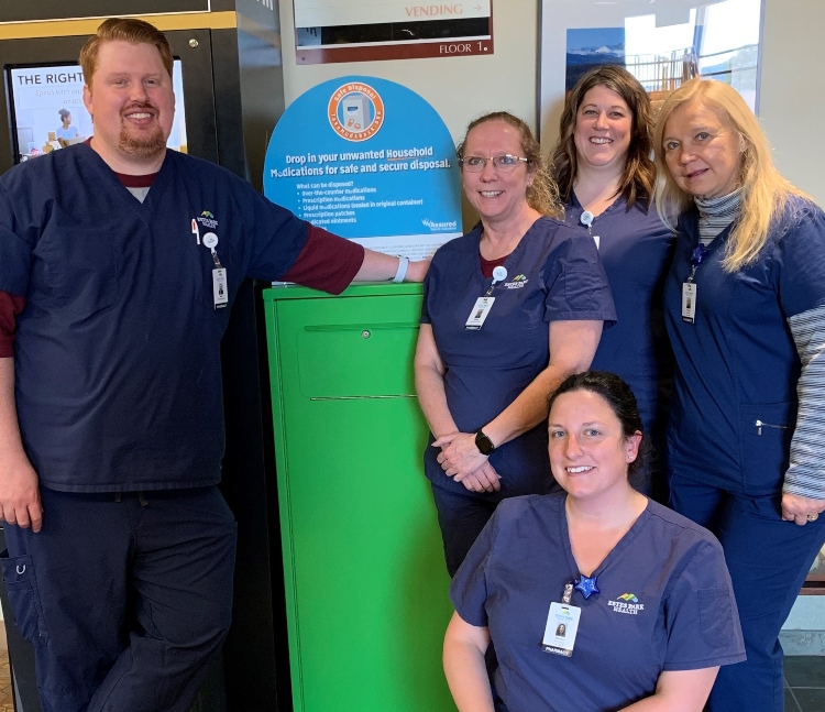 Five healthcare workers in navy scrubs stand and sit around a green medication disposal box, smiling at the camera inside a facility.