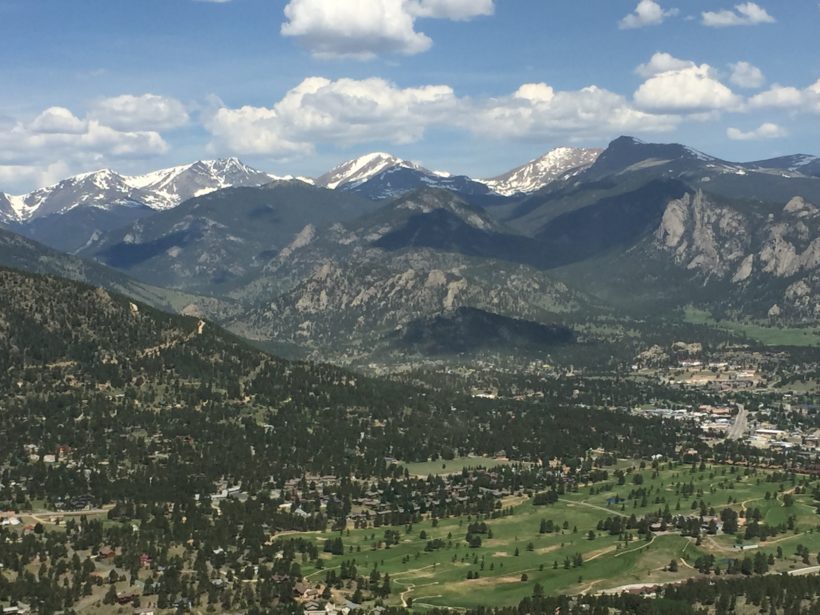 Aerial view of a mountainous landscape featuring a town nestled in a valley, with green fields and forested areas, under a partly cloudy sky. Snow-capped mountains are visible in the background.