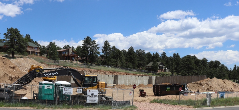 Construction site with excavators, portable toilets, and large containers. Signs indicate the presence of a construction company. Trees and homes are visible in the background under a partly cloudy sky.