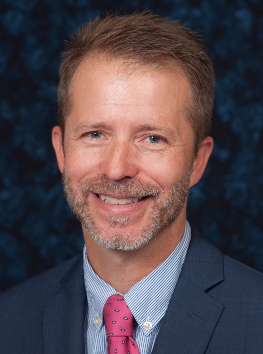 A man with short hair and a beard smiles at the camera. He is wearing a blue suit, a light blue shirt, and a pink tie with blue dots.