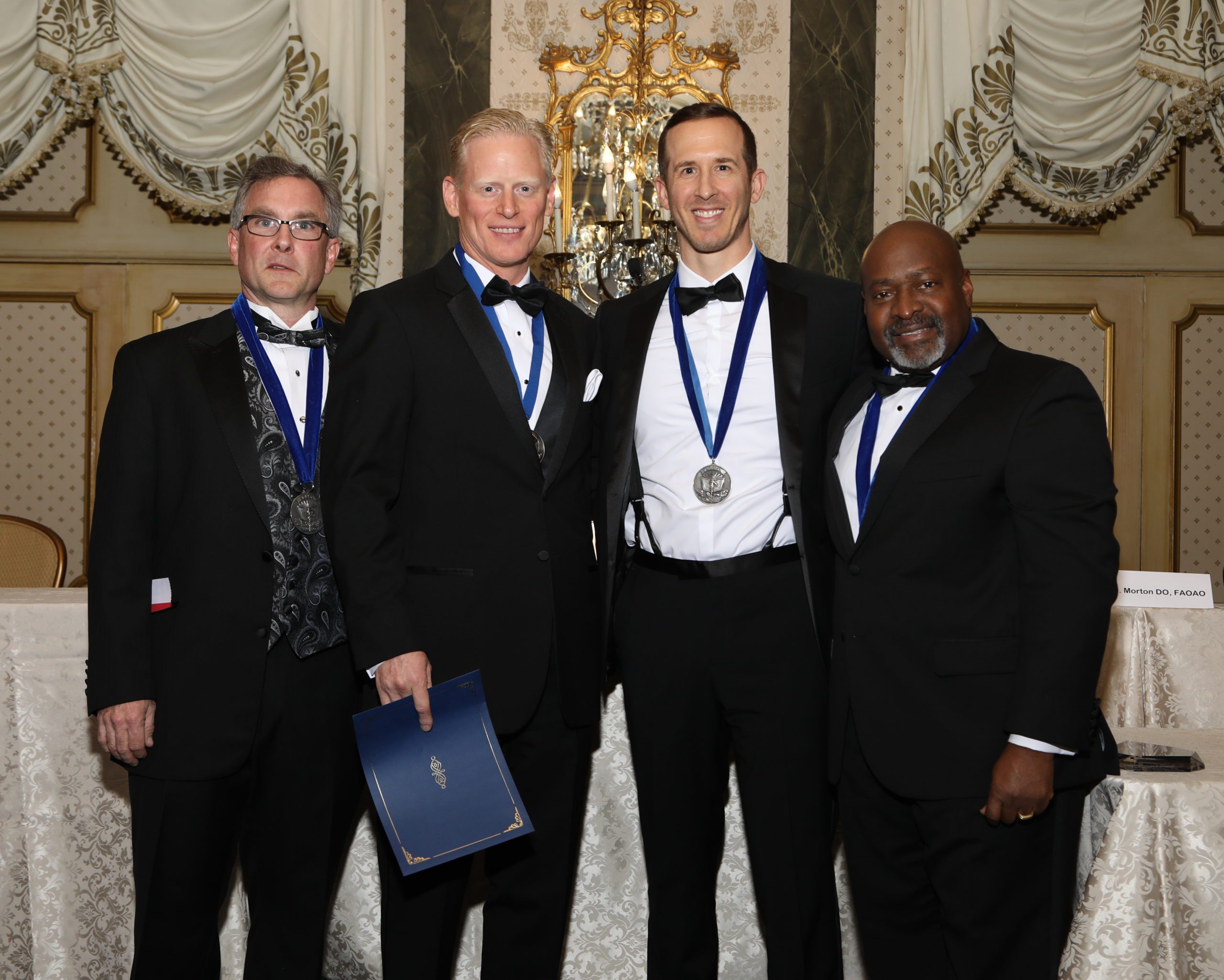 Four men in formal attire stand together in front of an ornate backdrop, three wearing medals. One man holds a folder, and they are posing for a photo at what appears to be an award ceremony.