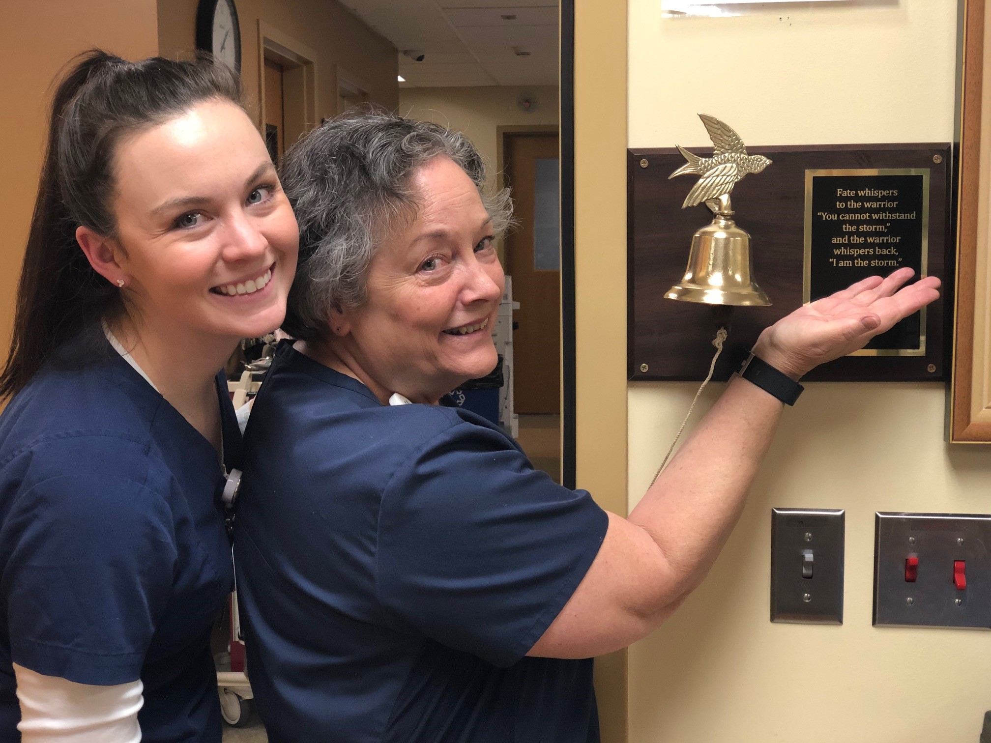 Two medical professionals in navy scrubs smile, with one ringing a brass bell mounted on a plaque. The plaque has an inscription next to it.