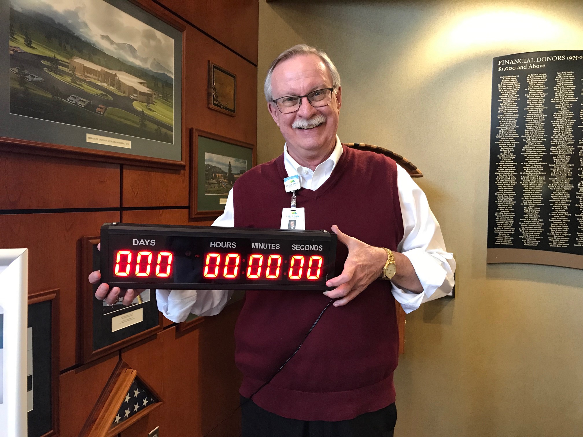 A man wearing glasses and a red vest smiles while holding a countdown clock showing all zeros. He stands in an office next to a wall with framed pictures and a donor recognition plaque.