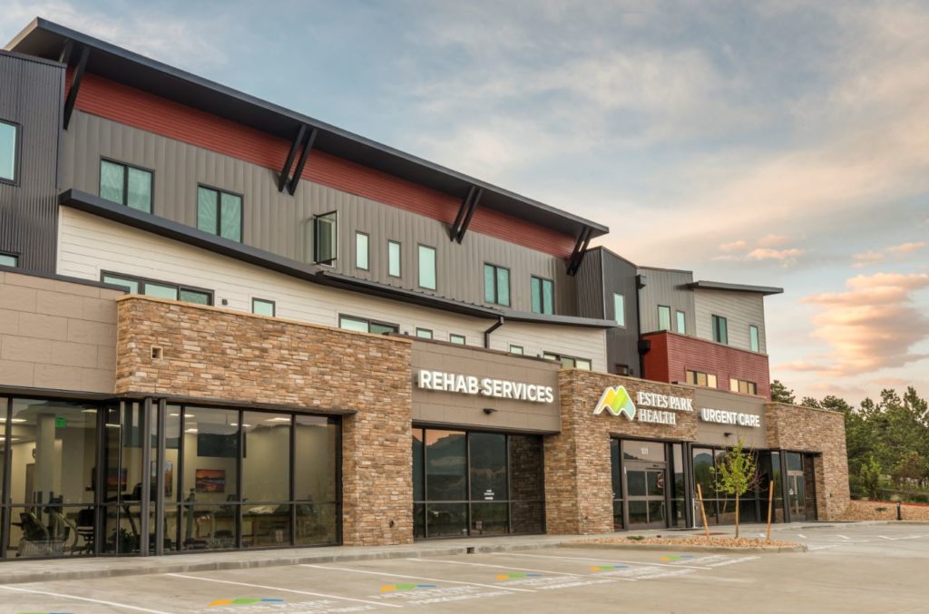 A modern, multi-story building houses "Rehab Services" and "Urgent Care" facilities, featuring a mix of brick and metal siding with a sunset sky in the background.