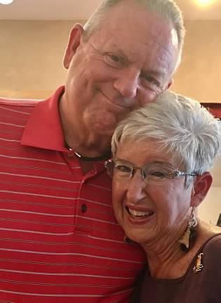 An elderly couple poses closely together, smiling warmly. The man wears a red striped polo shirt, and the woman wears glasses with short, gray hair.