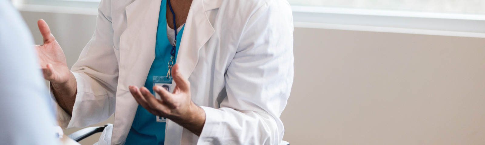 A healthcare professional wearing a white coat and blue scrubs gestures with their hands while speaking to someone across from them in a well-lit room.