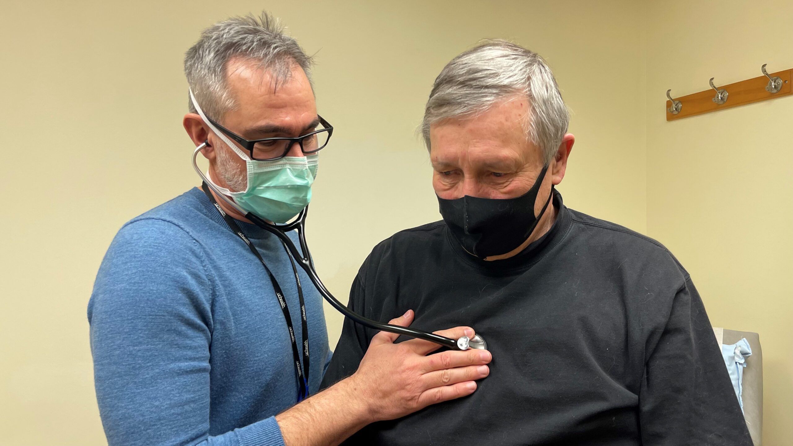 A healthcare professional uses a stethoscope to examine an elderly man, both are wearing face masks.