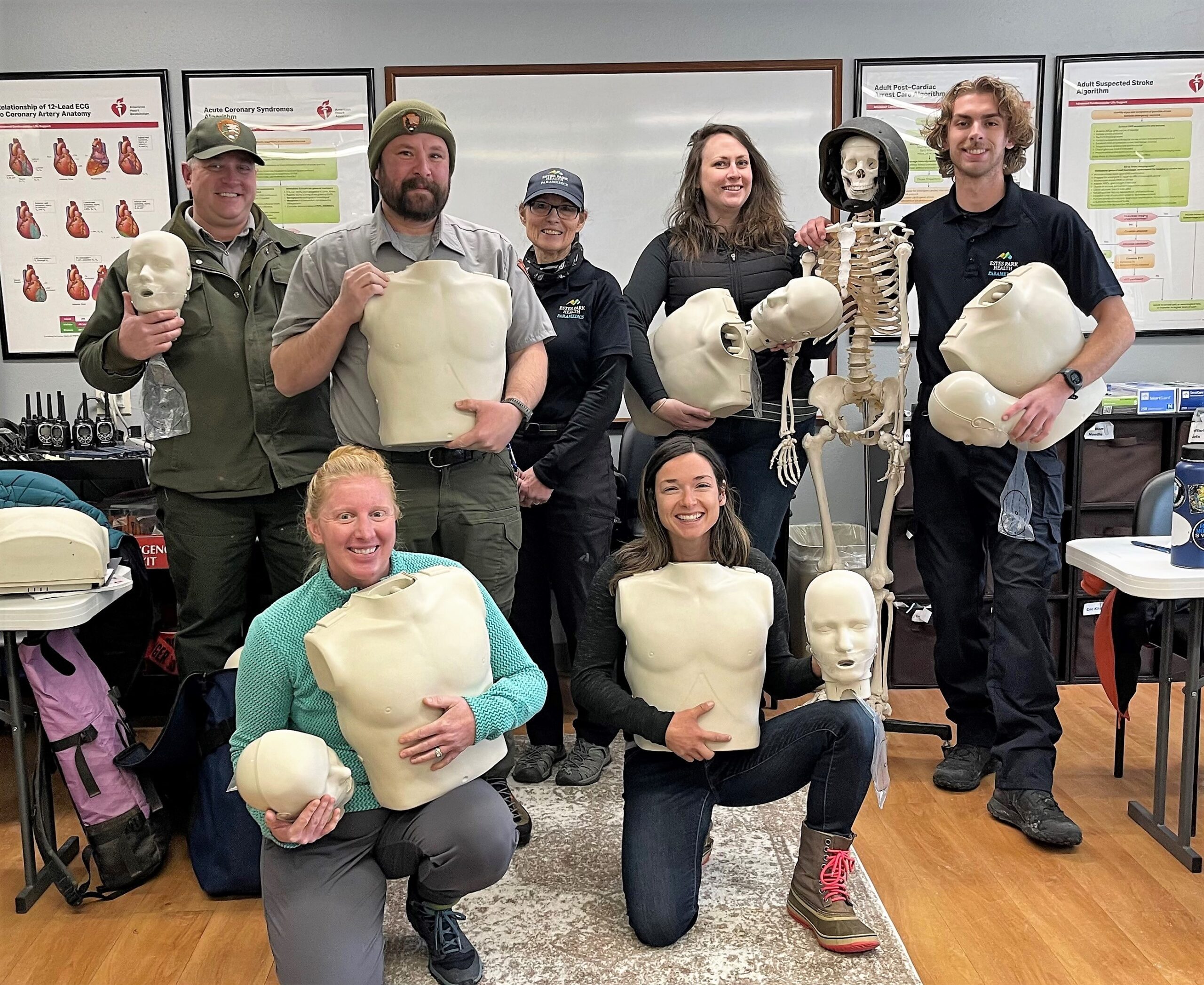 A group of seven people, holding CPR mannequins, poses in a classroom setting with educational posters on the walls and a skeleton model. There are CPR training materials in the background.