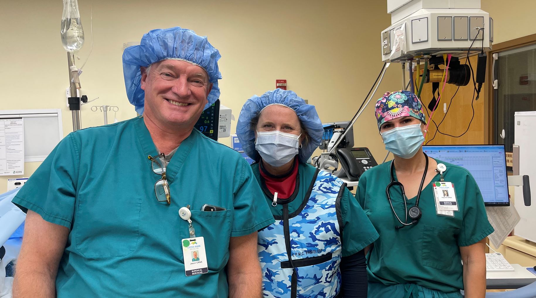 Three medical professionals wearing scrubs, face masks, and surgical caps stand in a hospital operating room, surrounded by medical equipment.