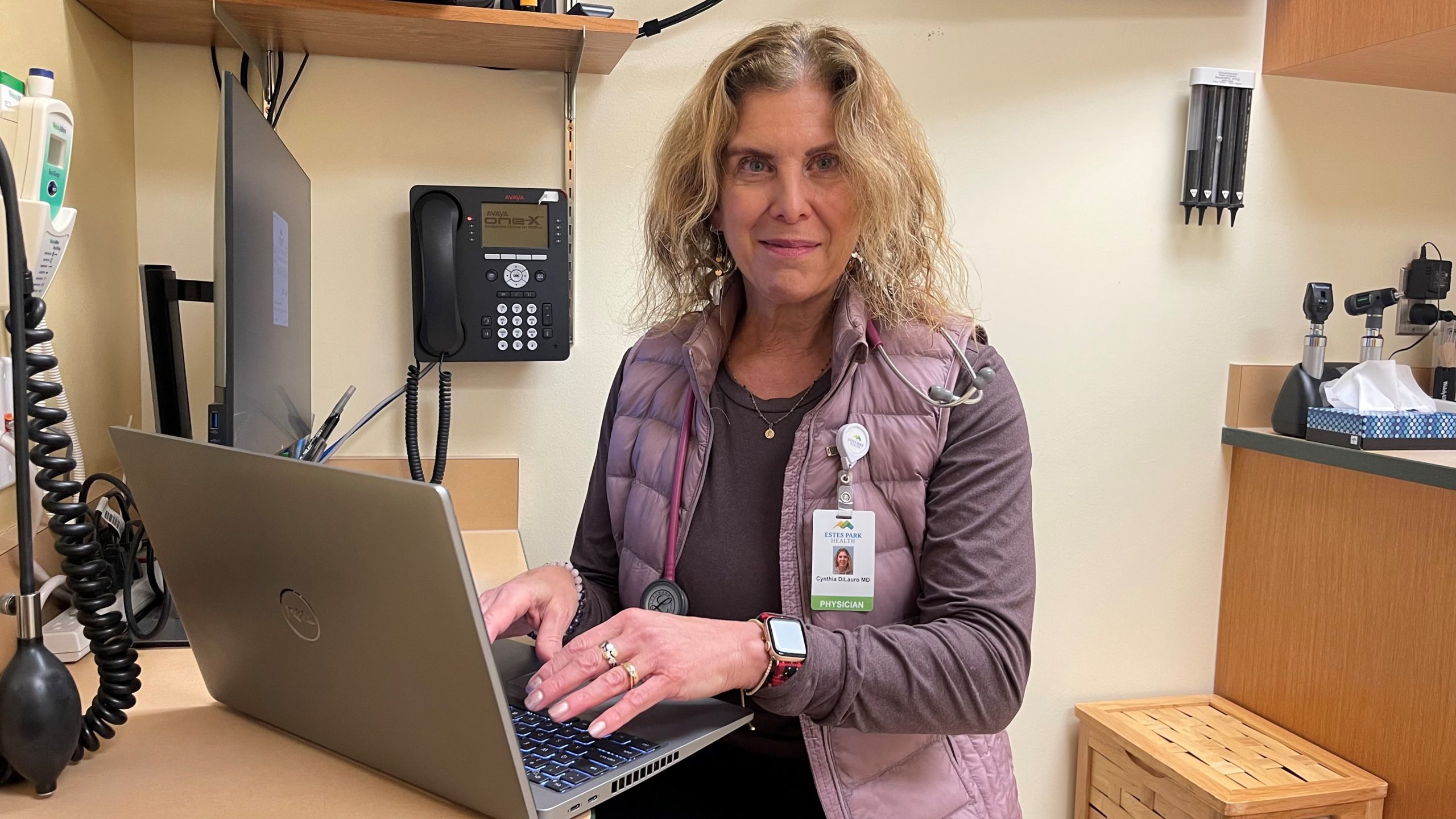 A woman in a medical setting is typing on a laptop. She is wearing a stethoscope around her neck, a work ID badge, and a smartwatch. Medical equipment can be seen in the background.