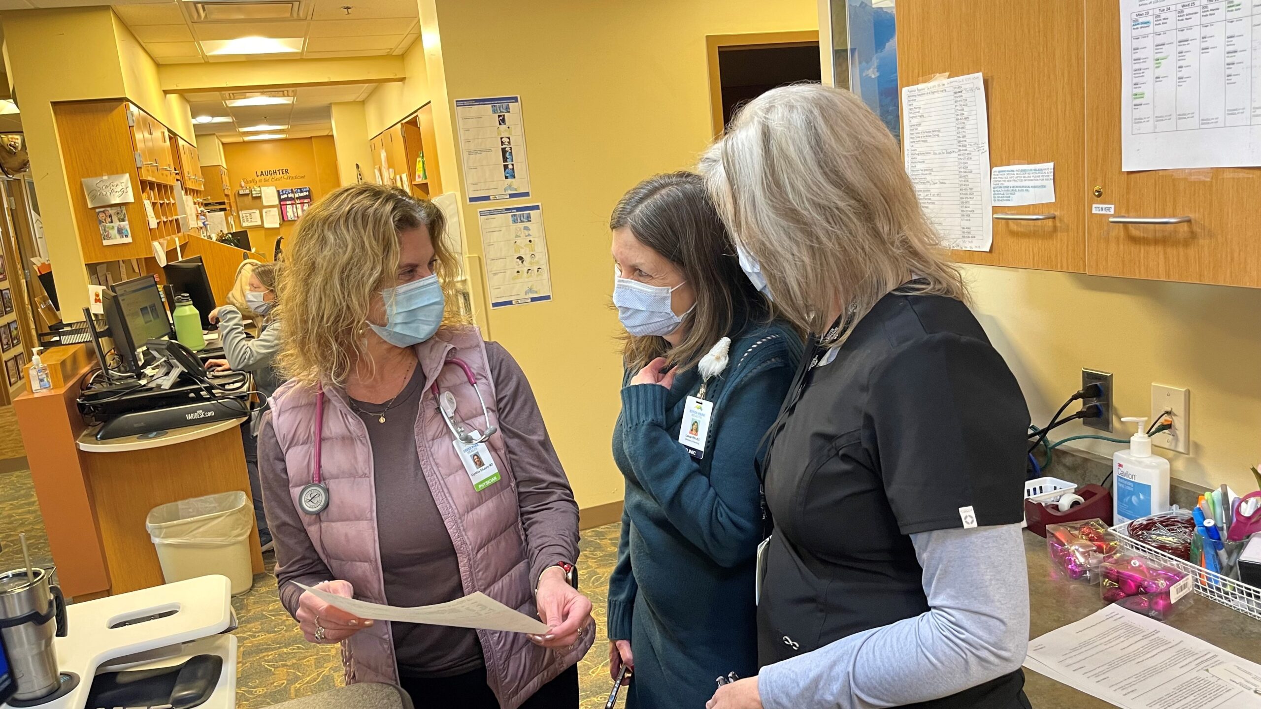 Three healthcare workers wearing masks discuss a document in a clinic setting, with office and medical supplies visible in the background.