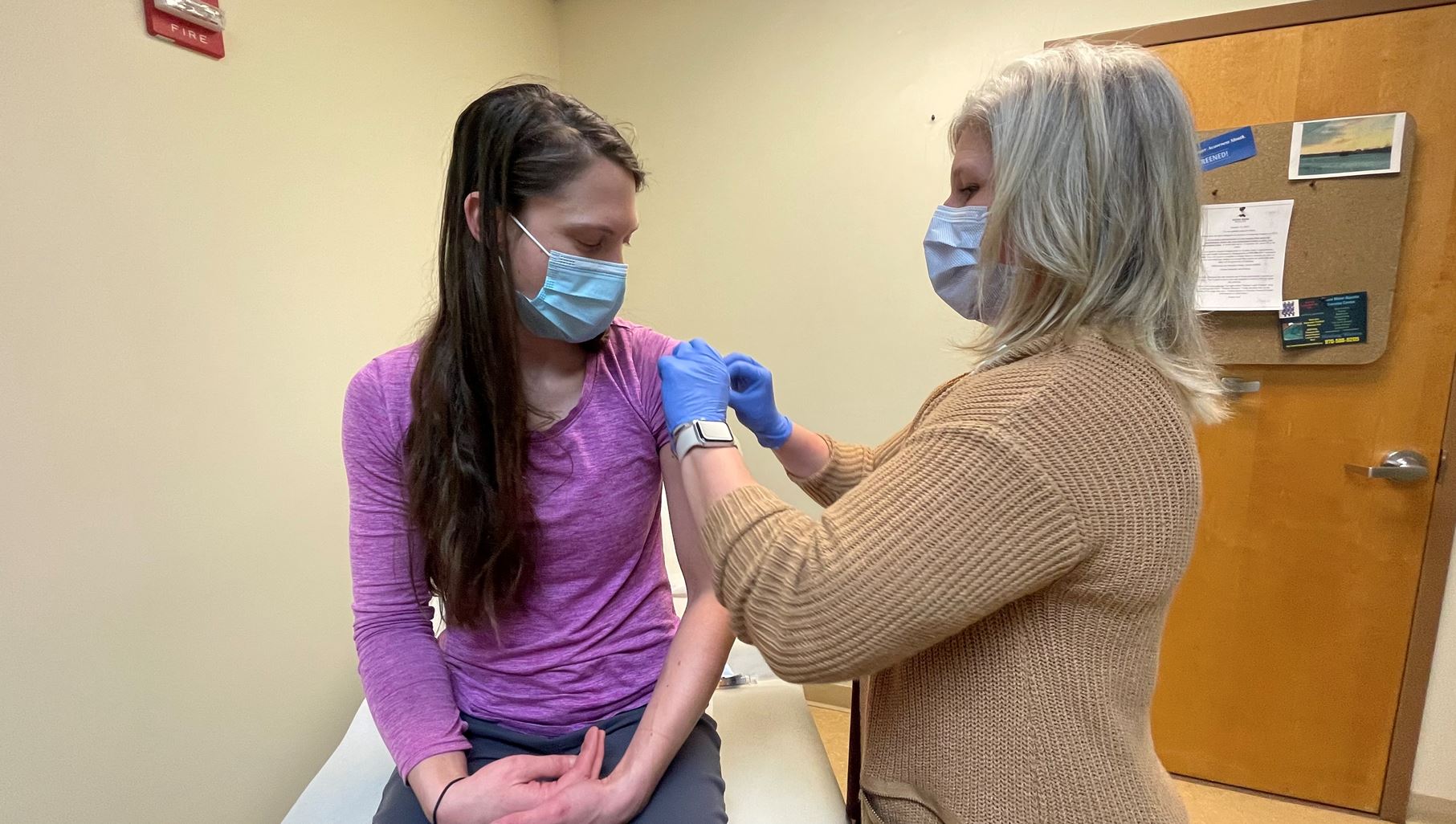 A person wearing a face mask and a purple shirt receives a vaccination from a healthcare worker in a medical setting.