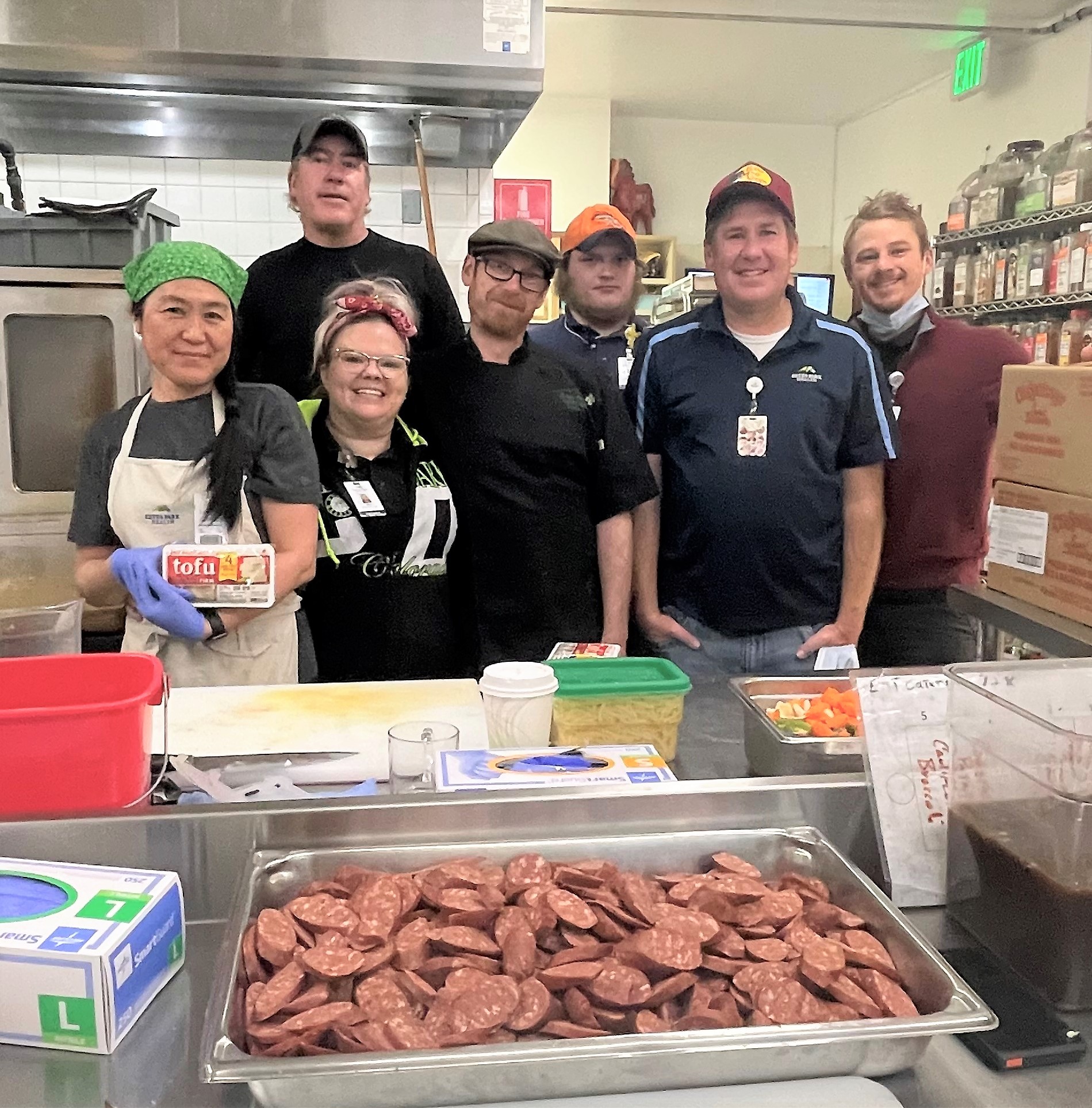 Seven people stand in a kitchen, four in aprons, posing for a group photo behind trays of sliced sausages and other ingredients.