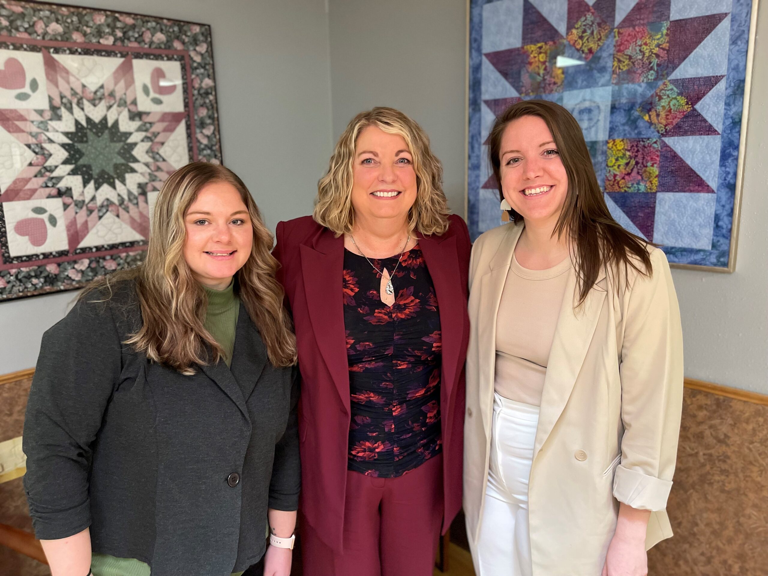 Three women stand side by side, smiling, in front of two quilted wall hangings. One is wearing a dark green top and gray jacket, another in a maroon suit, and the third in a beige jacket and tan top.