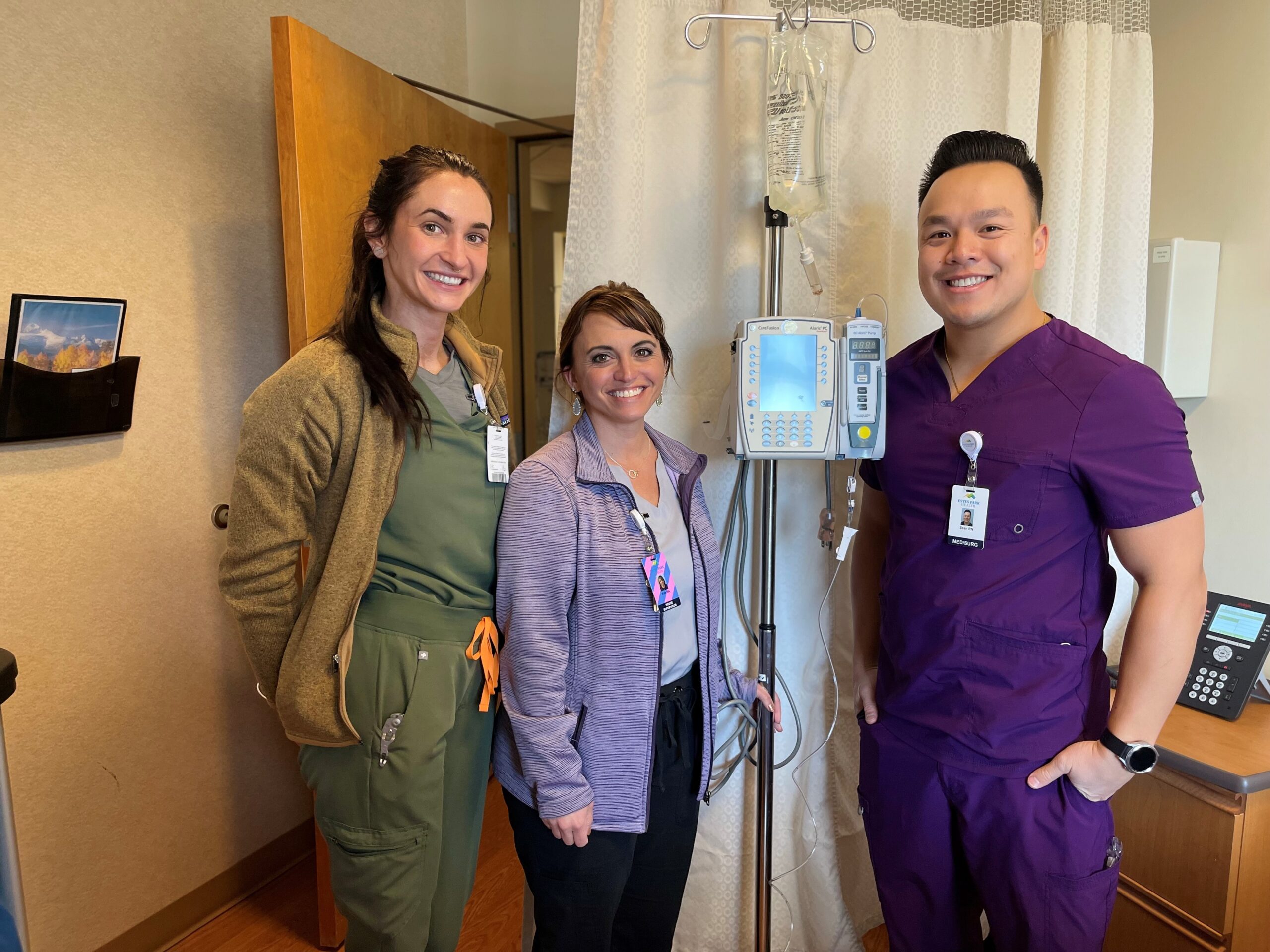 Three healthcare professionals in scrubs stand smiling next to medical equipment in a hospital room.