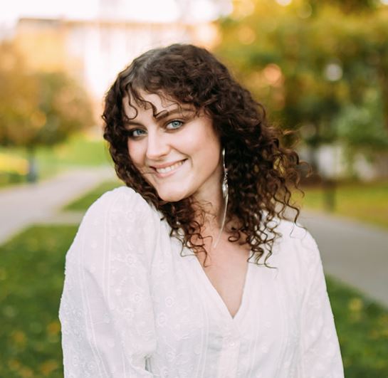 A person with curly hair and wearing a white top is smiling outdoors with blurred greenery in the background.