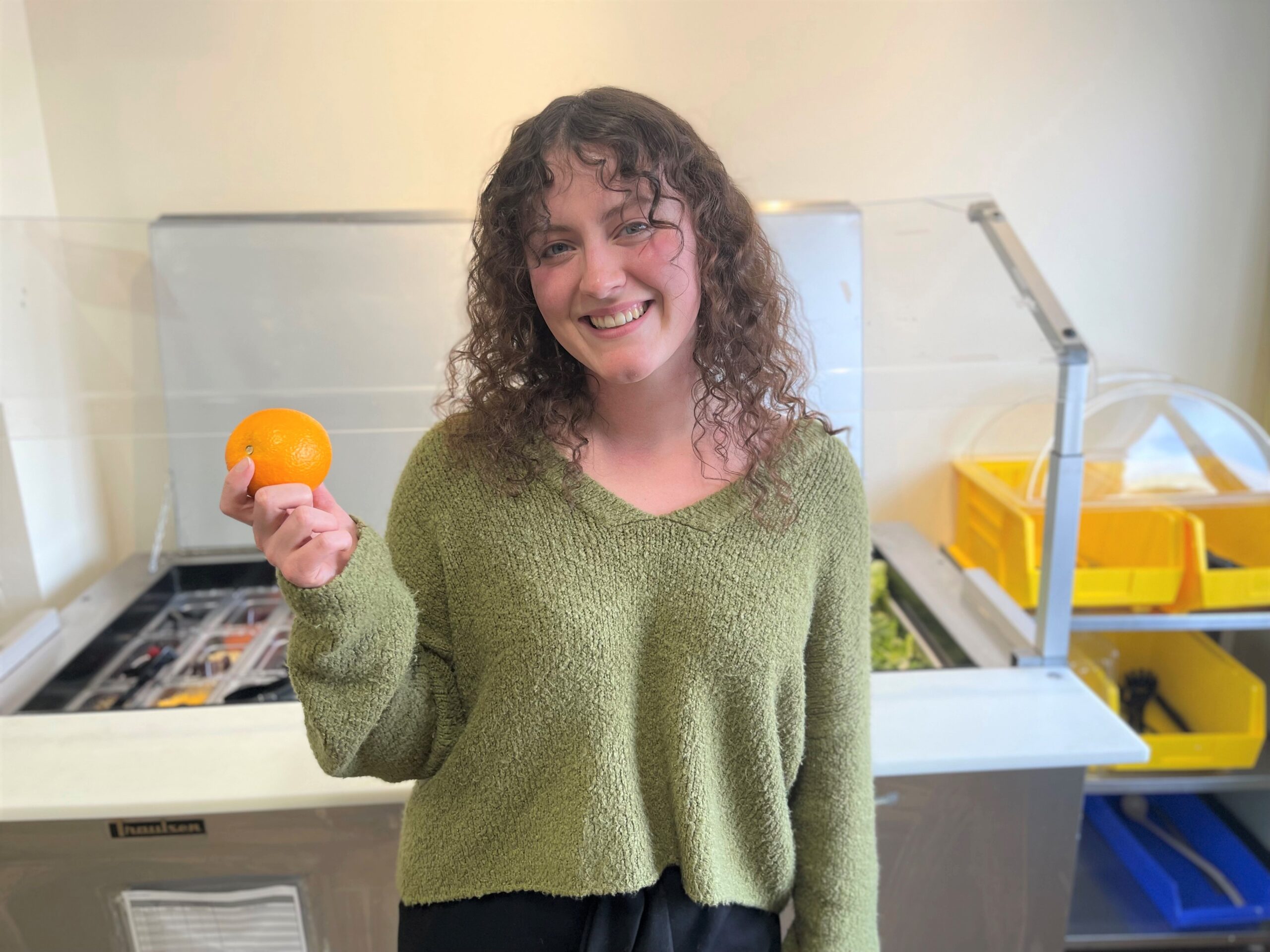 A person with curly hair and wearing a green sweater is smiling and holding an orange in their hand, standing in front of a food counter.