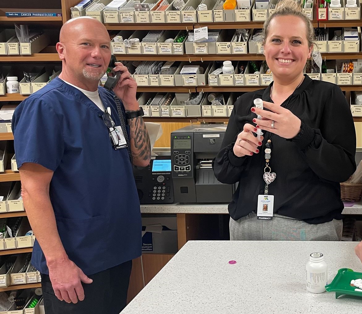 Two pharmacy staff members, one in blue scrubs and the other in black attire, stand behind a counter. The person in scrubs is on the phone, while the other holds a medication vial. Shelves with supplies fill the background.