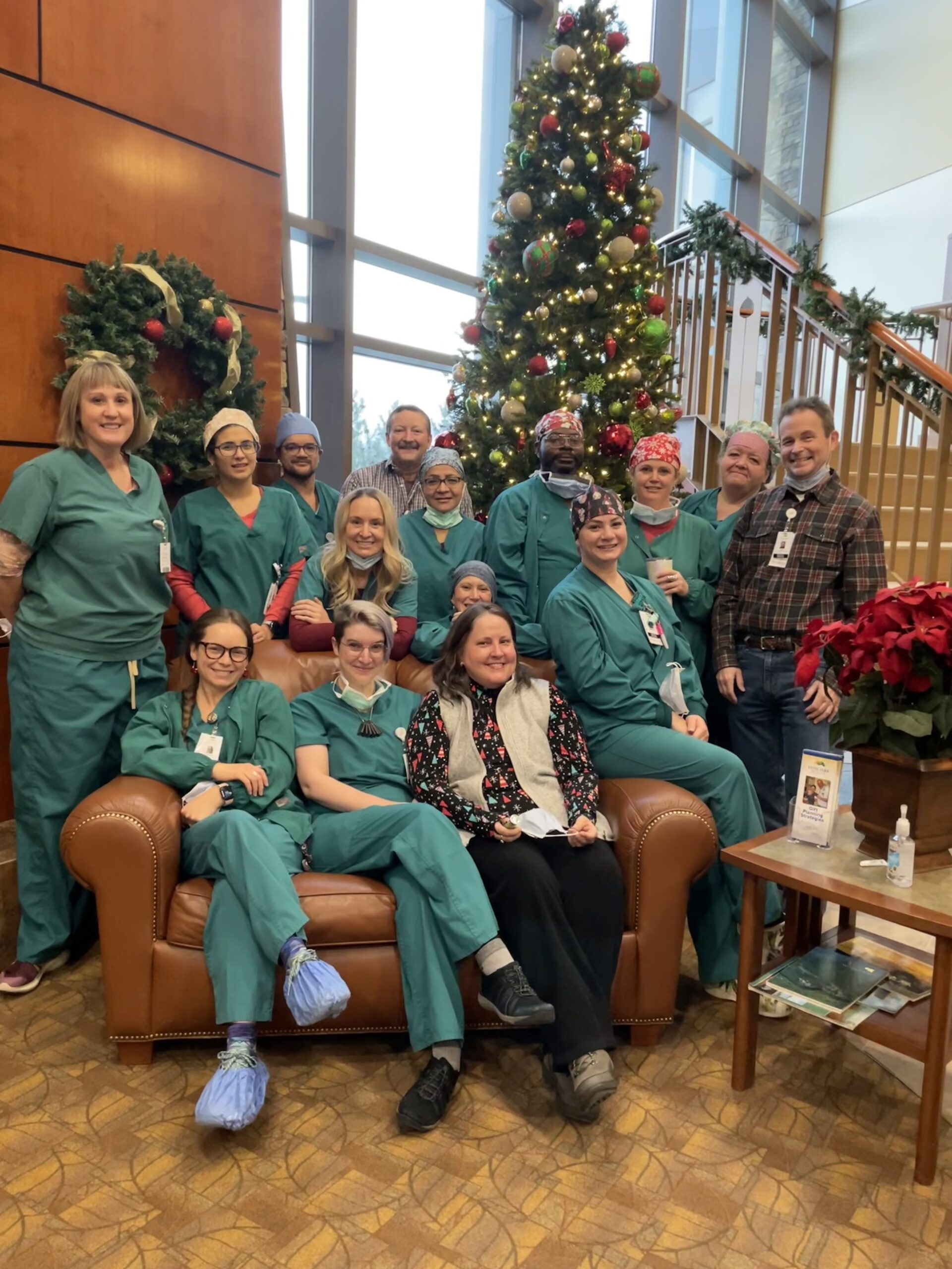 Group of healthcare professionals in green scrubs and a few staff in casual attire pose together in front of a decorated Christmas tree and wreath inside a building with large windows.