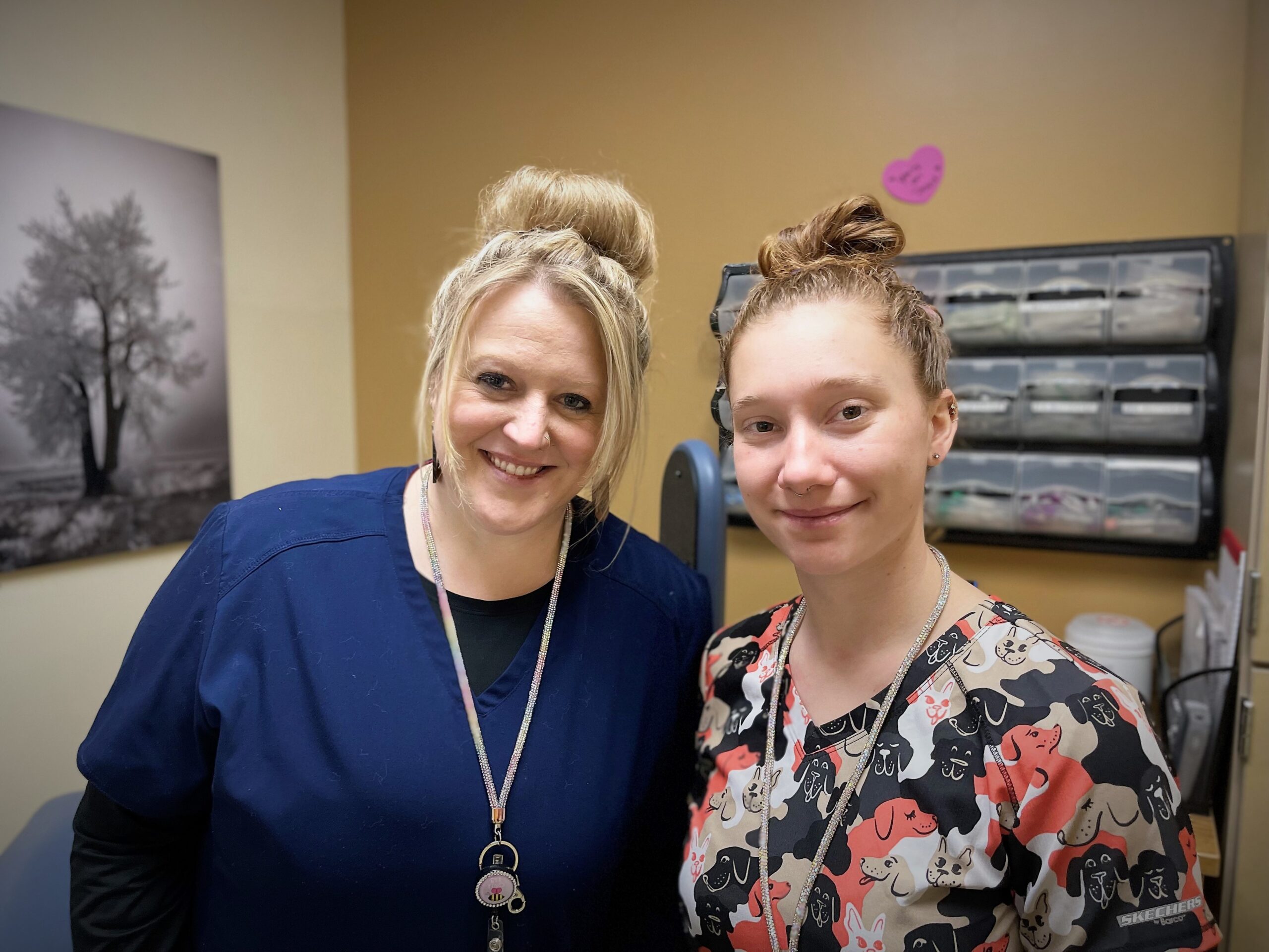 Two women standing side by side in a medical office. One wears navy scrubs and the other wears scrubs with a colorful dog print. A black and white tree photo is on the wall behind them.