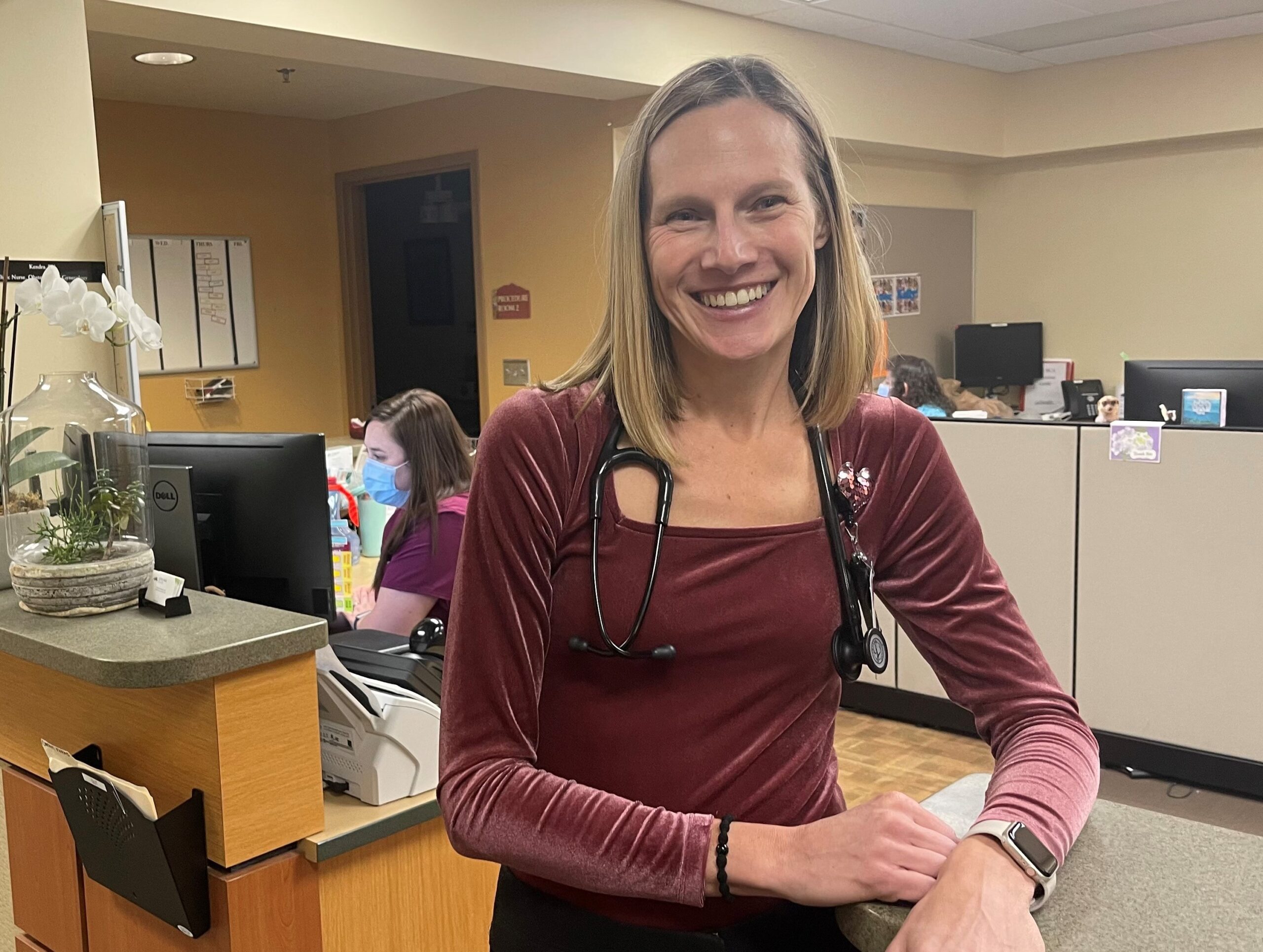 A woman wearing a stethoscope and a maroon top smiles while leaning on a counter in a medical office. Another person in purple scrubs works at a computer in the background.