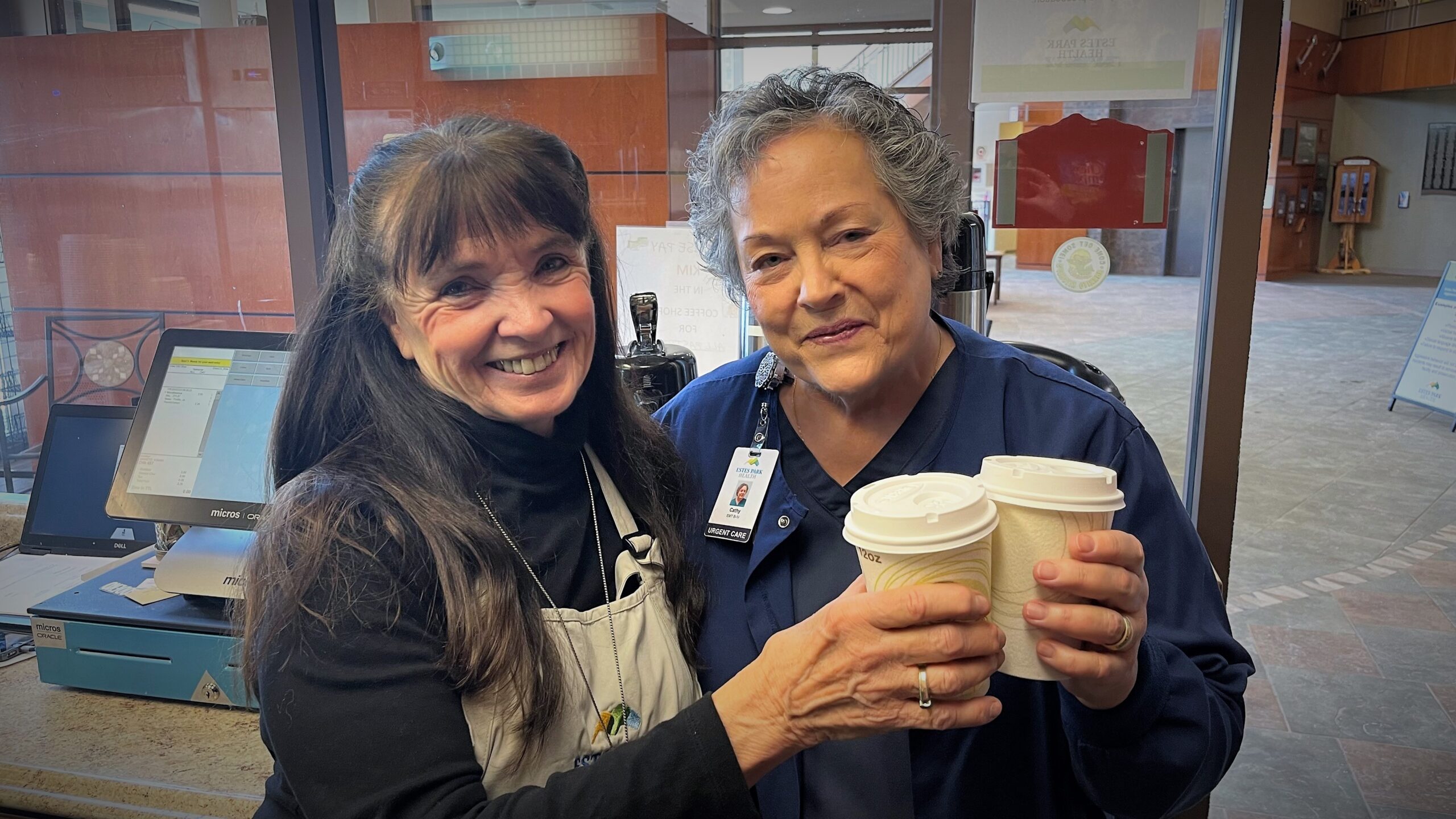 Two women, one in a black shirt and apron, and the other in a blue uniform, stand inside a building holding paper coffee cups. They are smiling and looking at the camera.