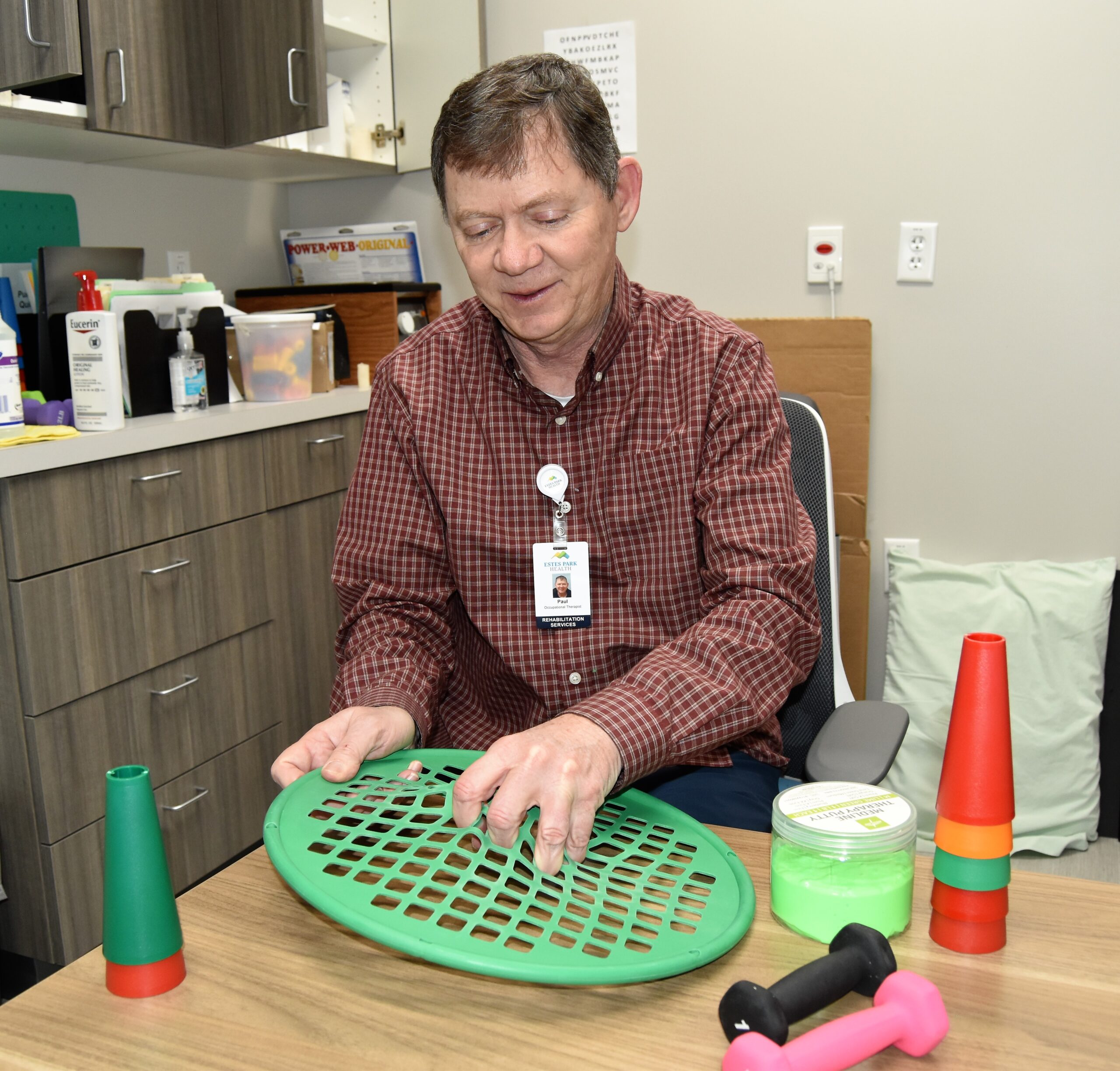 A man sits at a desk with physical therapy equipment, including cones, a green perforated disc, exercise bands, and small dumbbells. He is wearing a work ID badge and appears to be setting up.