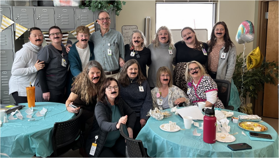 A group of people, all wearing fake mustaches, pose for a photo in a decorated room with tables and food items.