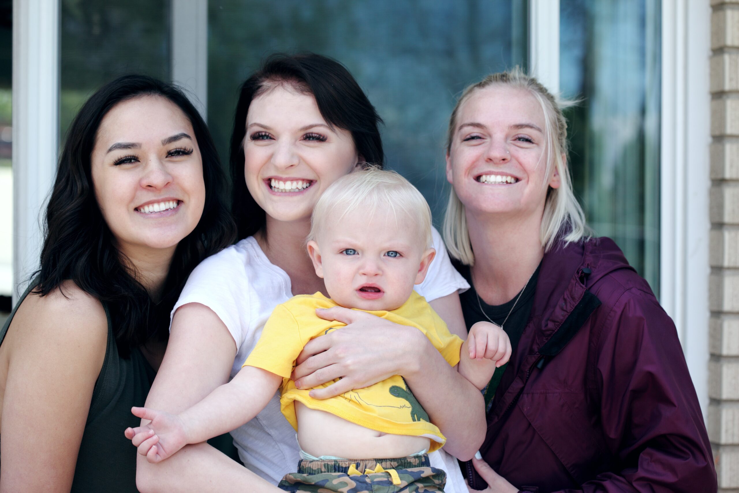 Three women smile at the camera while one holds a baby. They are standing outside in front of a window.