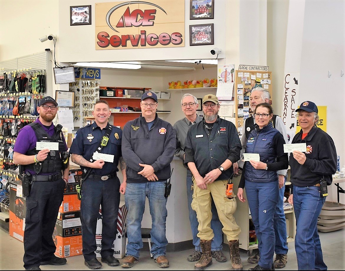 A group of eight people, including store staff and emergency responders, pose for a photo inside a hardware store, holding checks. A sign above the group reads 