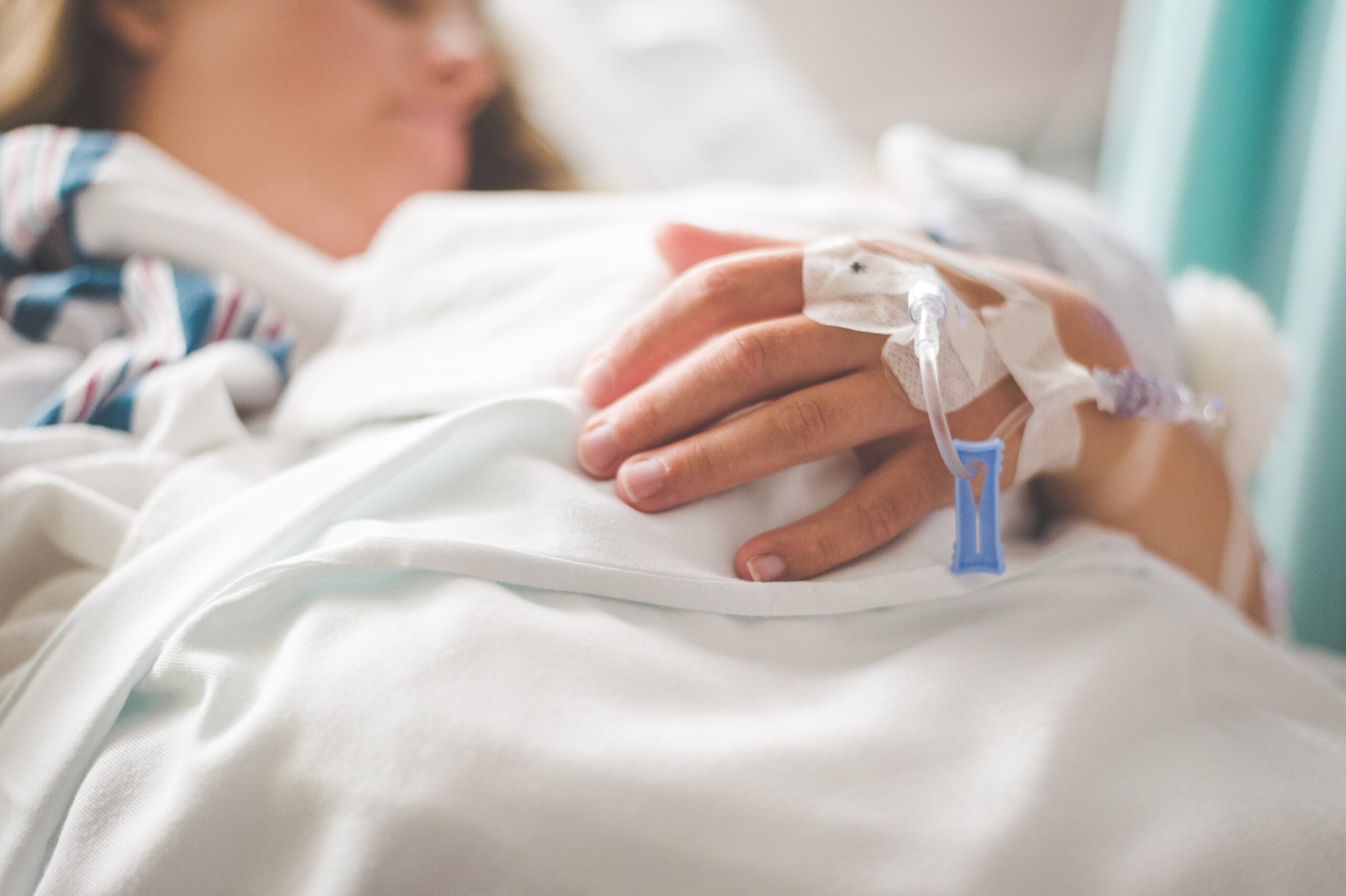 A person rests in a hospital bed with an intravenous line inserted in their hand.