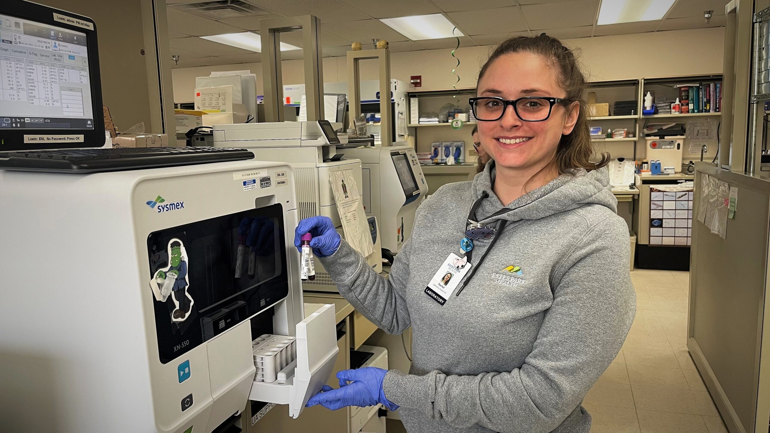 A lab worker in a gray hoodie and blue gloves uses a scientific instrument in a laboratory setting. She is holding a small vial and is smiling at the camera.