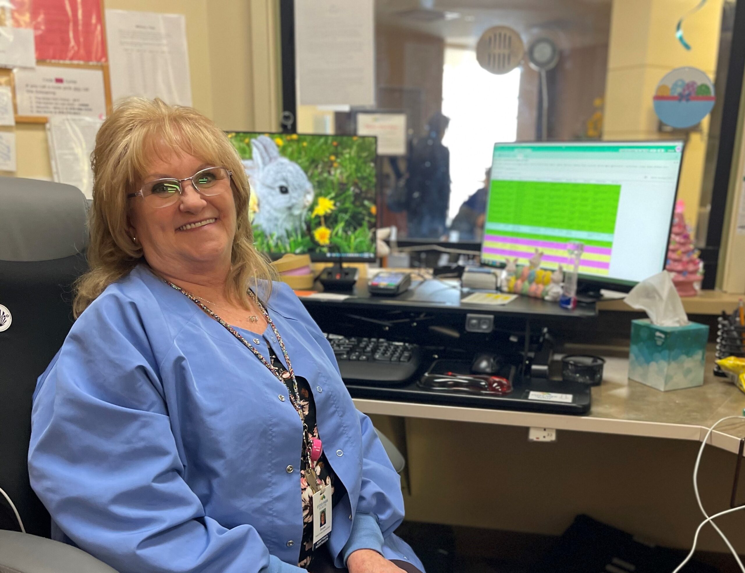 Smiling woman in a blue shirt sits at a desk with a dual monitor setup; one displays a rabbit, the other shows a spreadsheet. The desk holds various office supplies, a tissue box, and personal items.