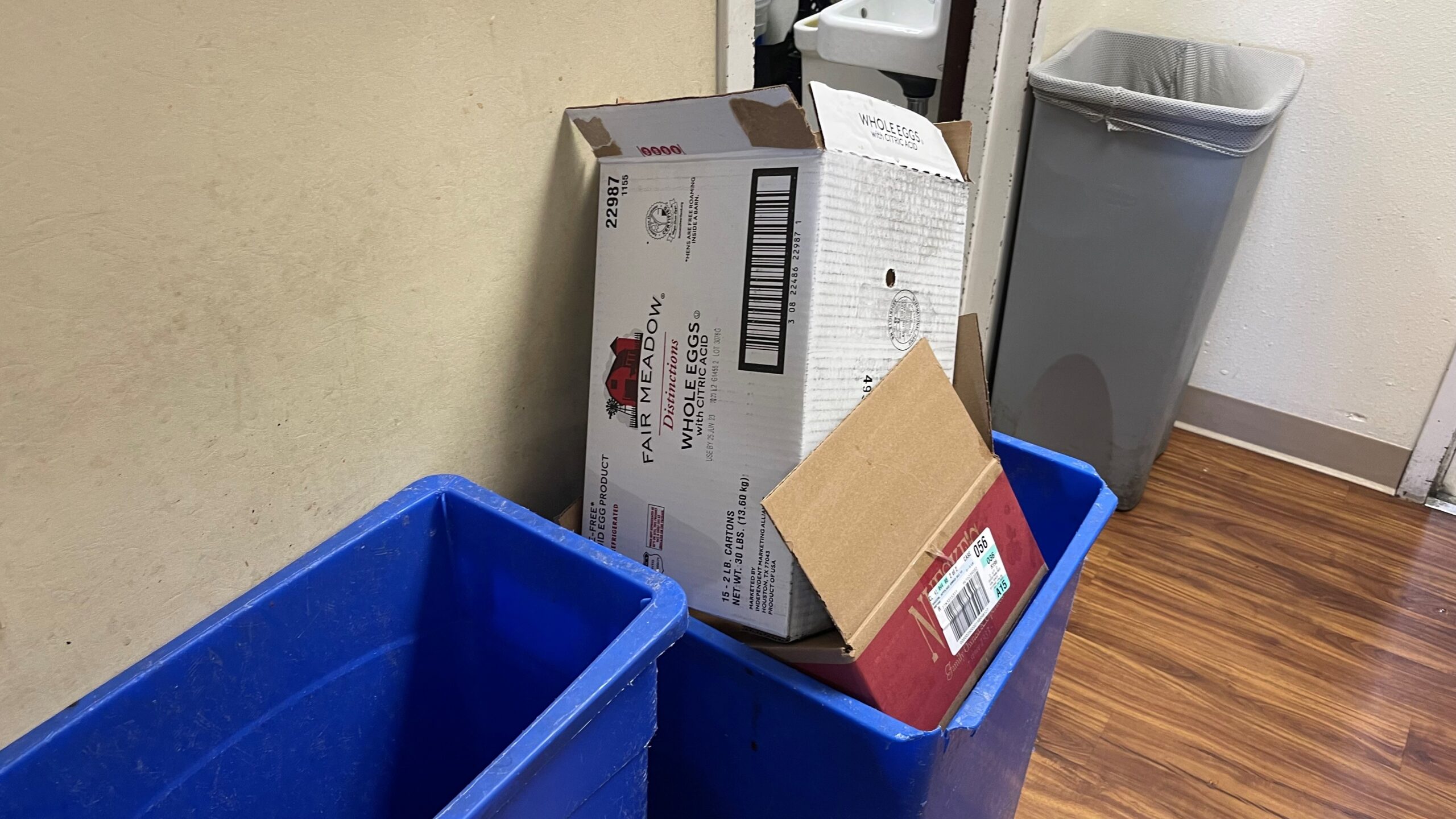 Cardboard boxes and a blue plastic bin are placed against a beige wall near a doorway and a grey trash can on a wooden floor.