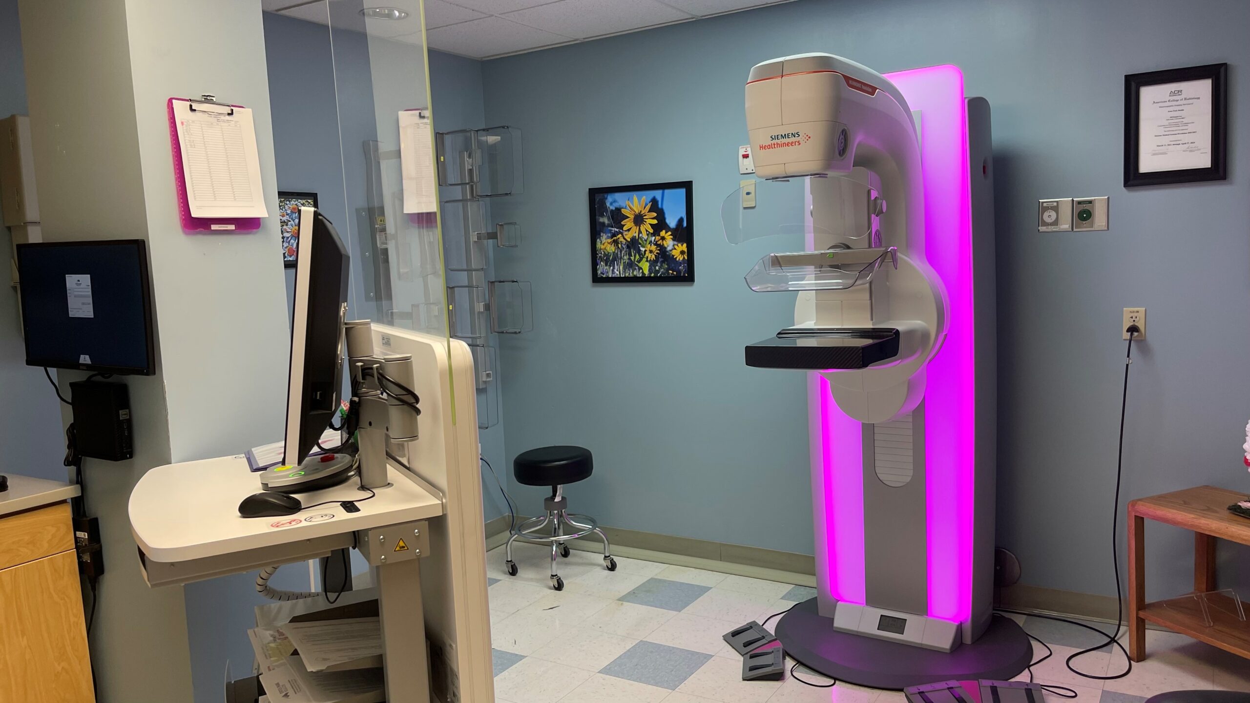 A mammography machine with a pink glow sits in a medical examination room with a computer workstation, wall art, and a certificate on the wall.