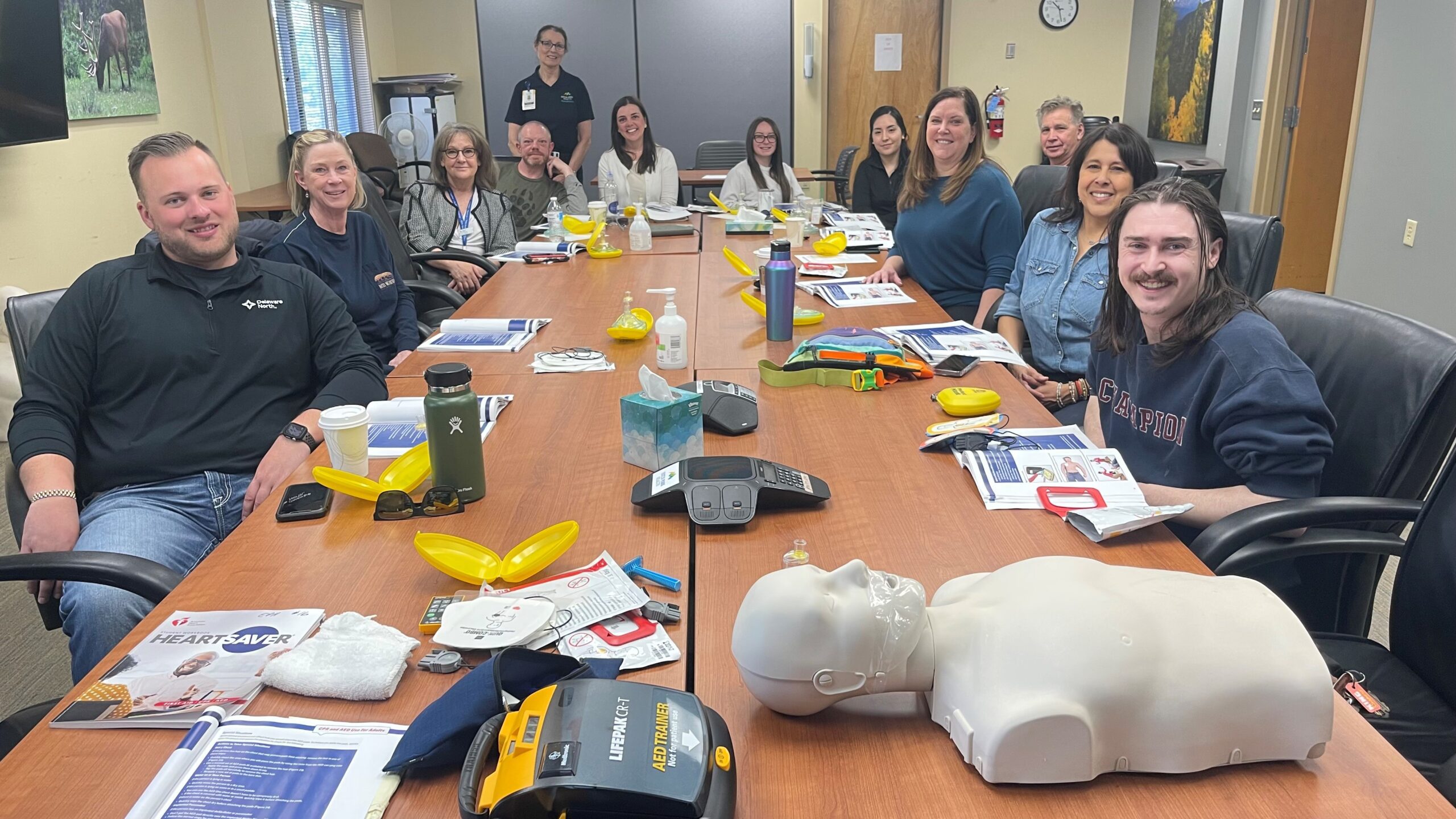 A group of people are seated around a conference table with CPR training materials, including a CPR mannequin, defibrillators, and instructional booklets.