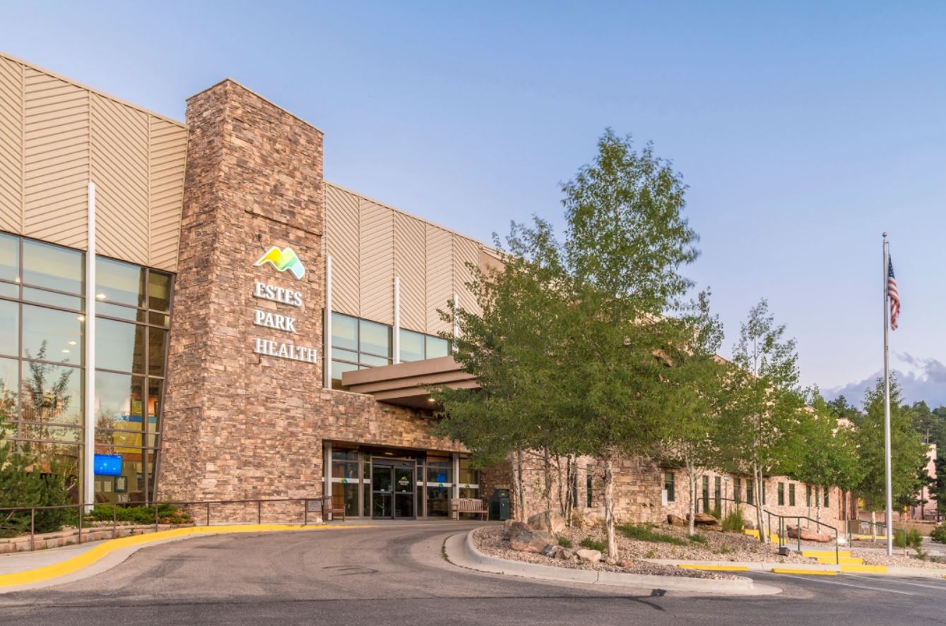 A wide view of the entrance to Estes Park Health, featuring a stone facade, large windows, and a pathway surrounded by landscaping, with a flagpole flying the American flag in the foreground.