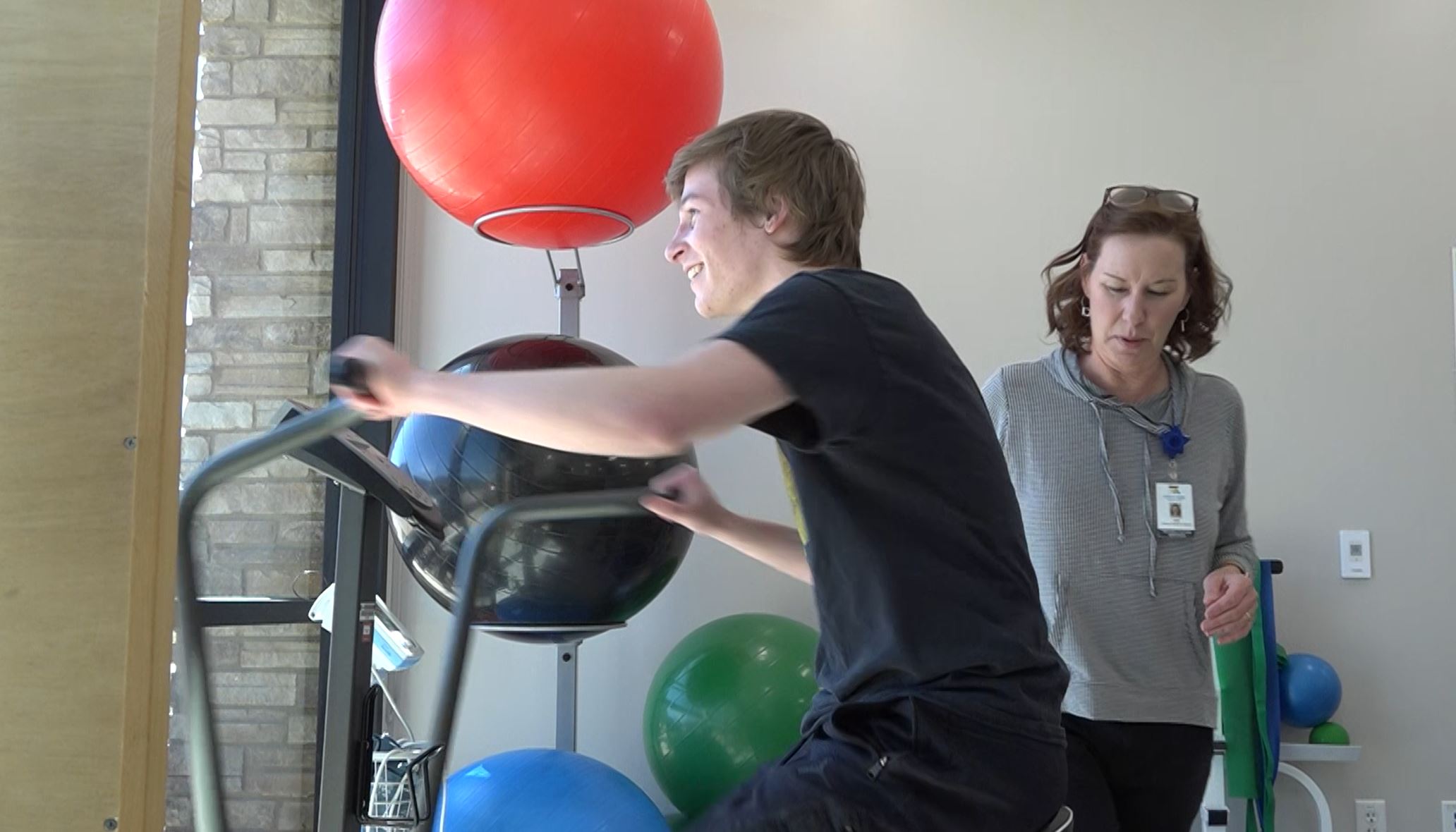 A young man exercises on a stationary bike while a woman, possibly a physical therapist, observes him, standing next to gym equipment and colorful exercise balls.