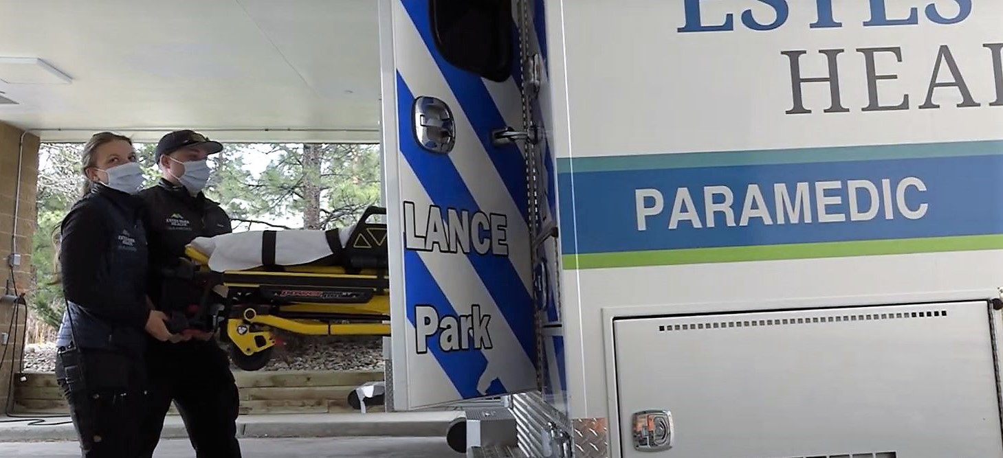 Two paramedics wearing masks load a patient on a stretcher into an ambulance marked 