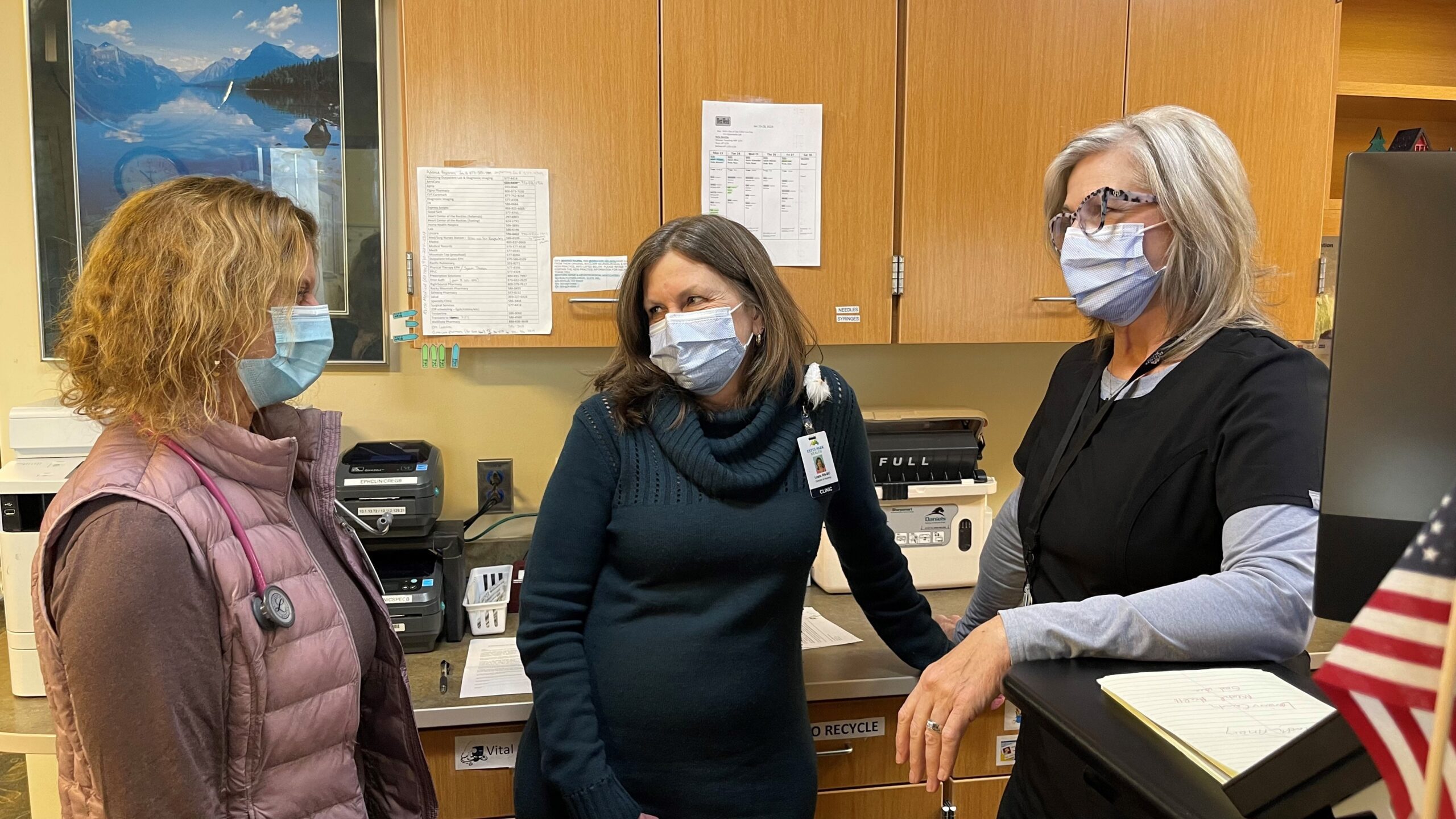 Three women wearing face masks are conversing in a medical office. One woman in a black uniform stands next to a computer, another in a green sweater hangs a badge, and the third wears a pink vest.