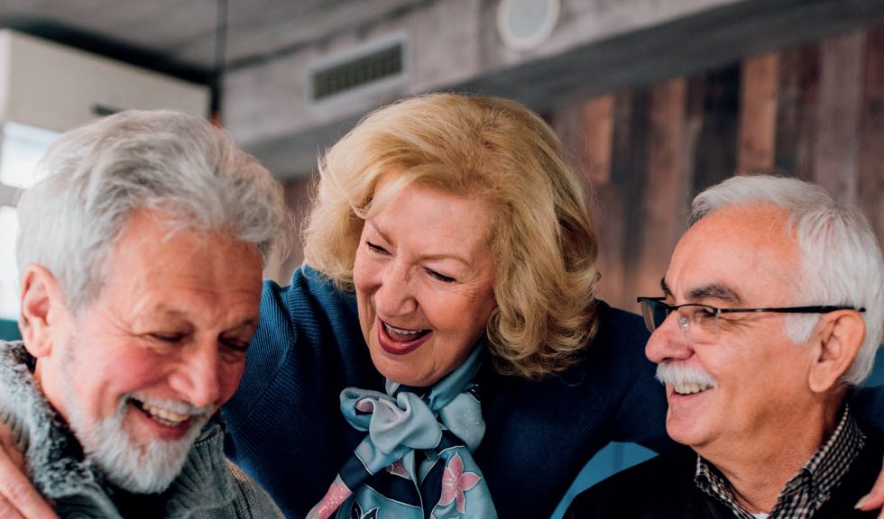 Three elderly individuals are shown socializing and smiling together. One person in the center has their arms around the others. The setting appears to be indoors.