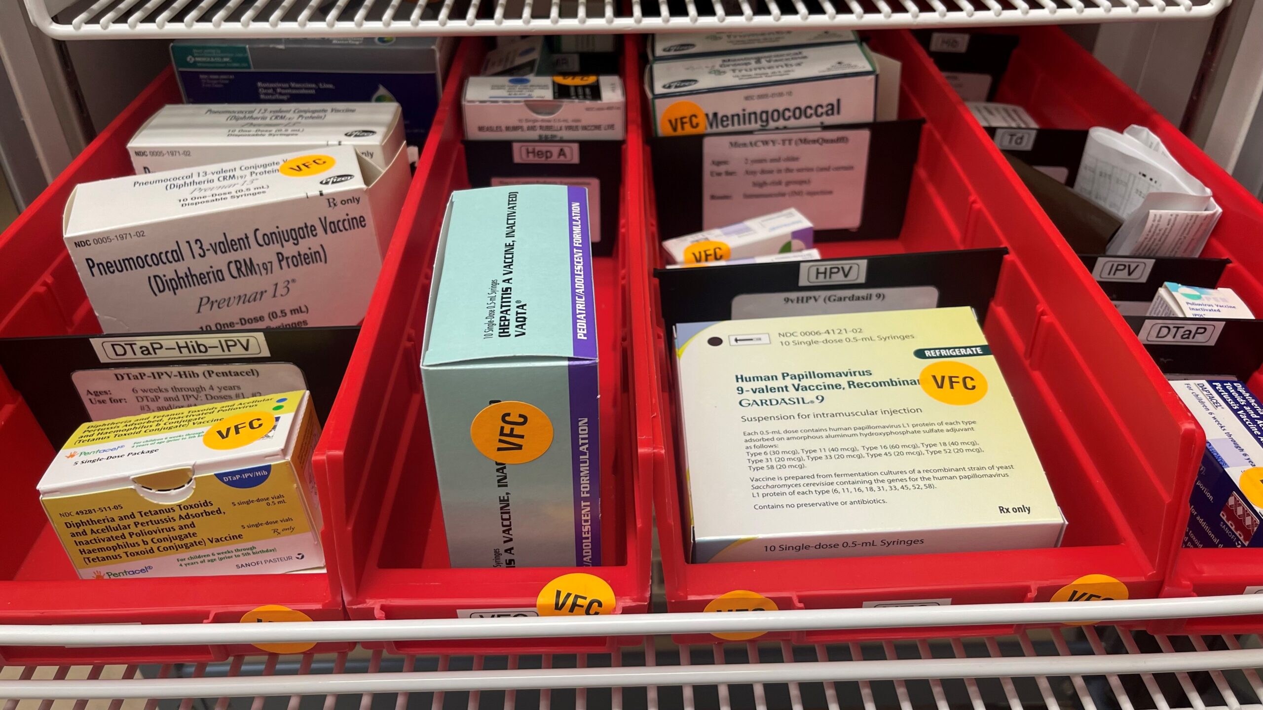 Red trays filled with various vaccine packages, including Pneumococcal, Meningococcal, HPV, and DTaP-IPV, labeled with VFC tags.