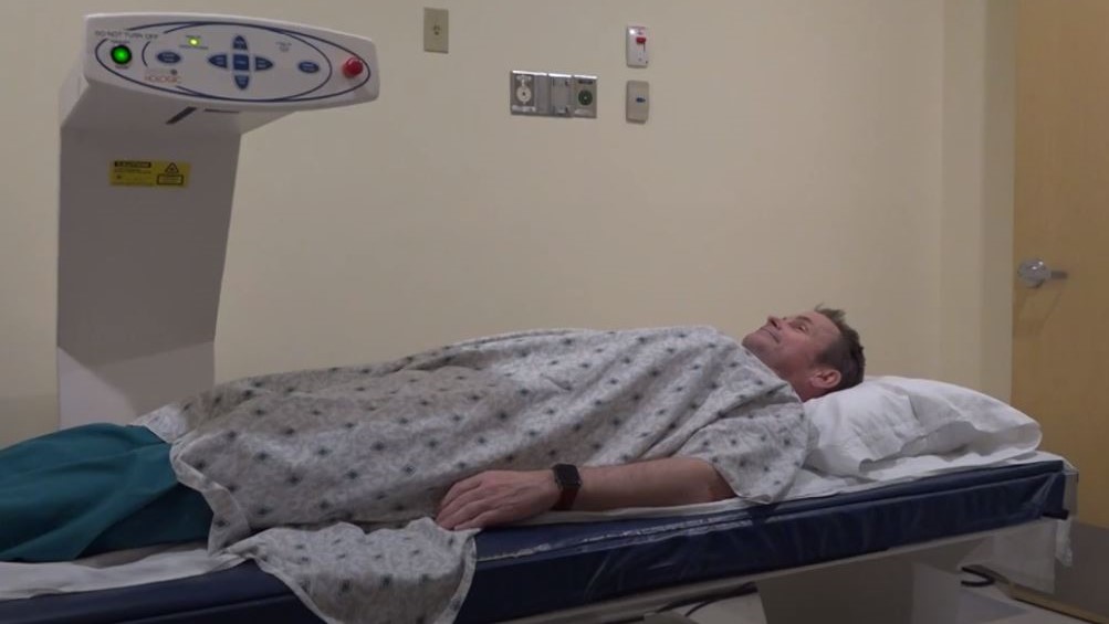 A person in a hospital gown lies on a medical examination table under a scanning device in a clinical room.