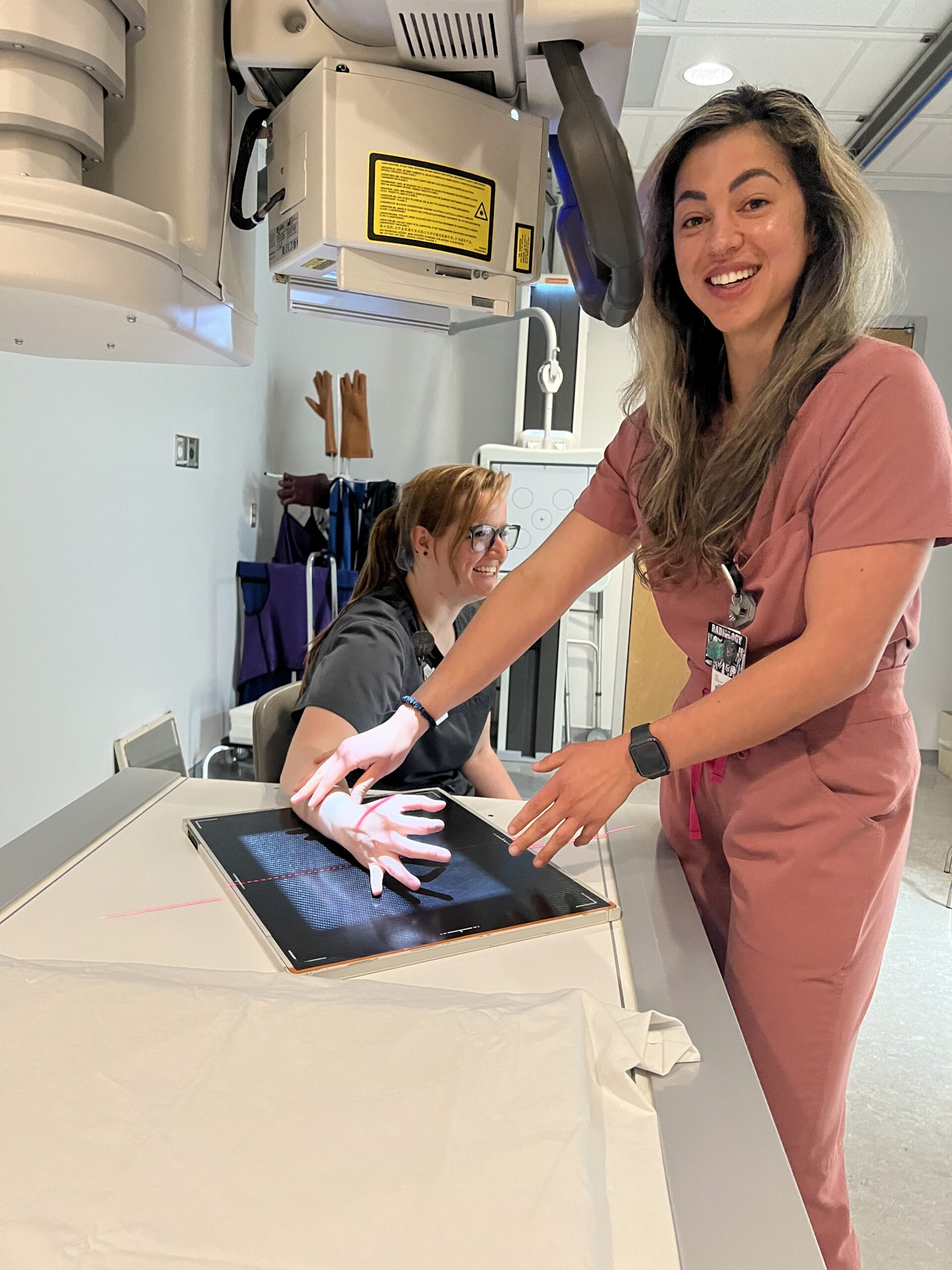 A person in pink scrubs stands smiling near an X-ray machine, pointing at an image of a hand on the screen, while another person in dark scrubs sits nearby.