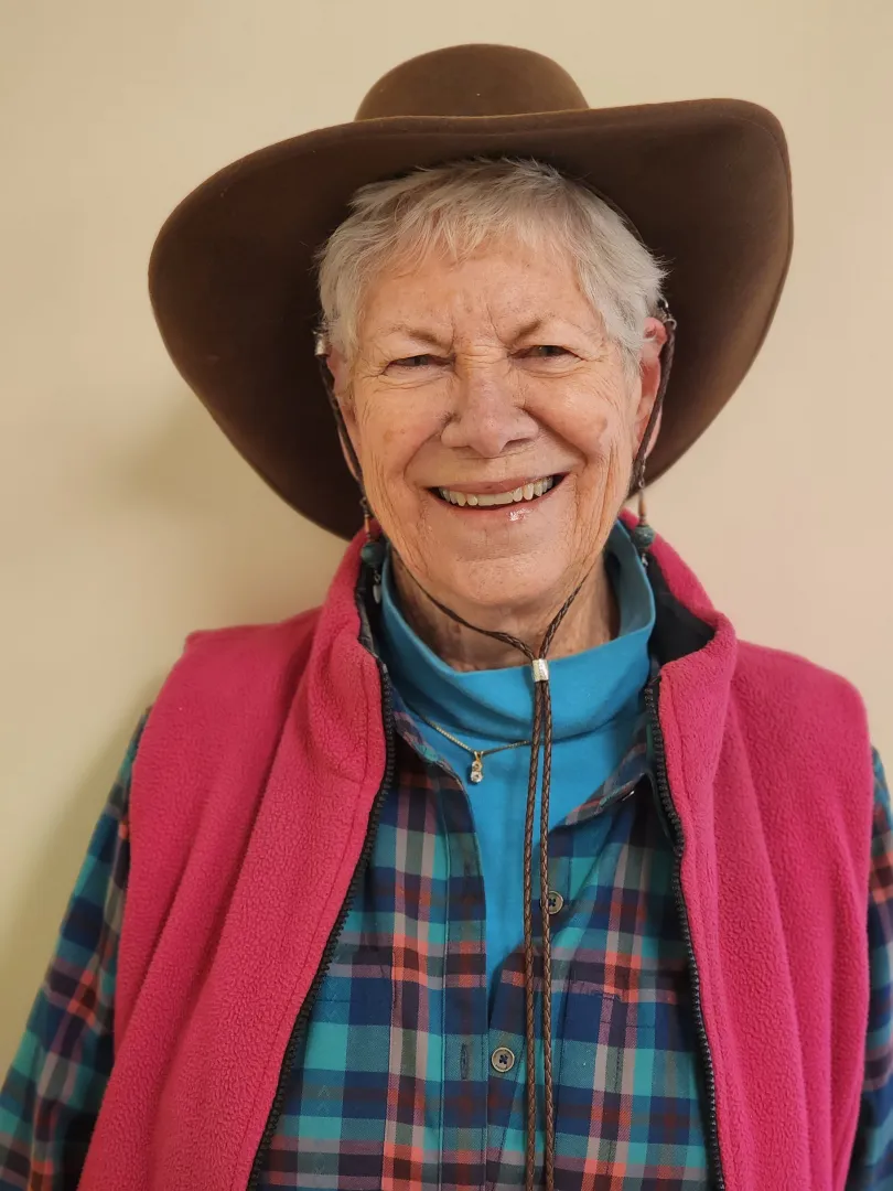 An elderly person smiling, wearing a brown cowboy hat, pink vest, and checked shirt, standing against a beige background.